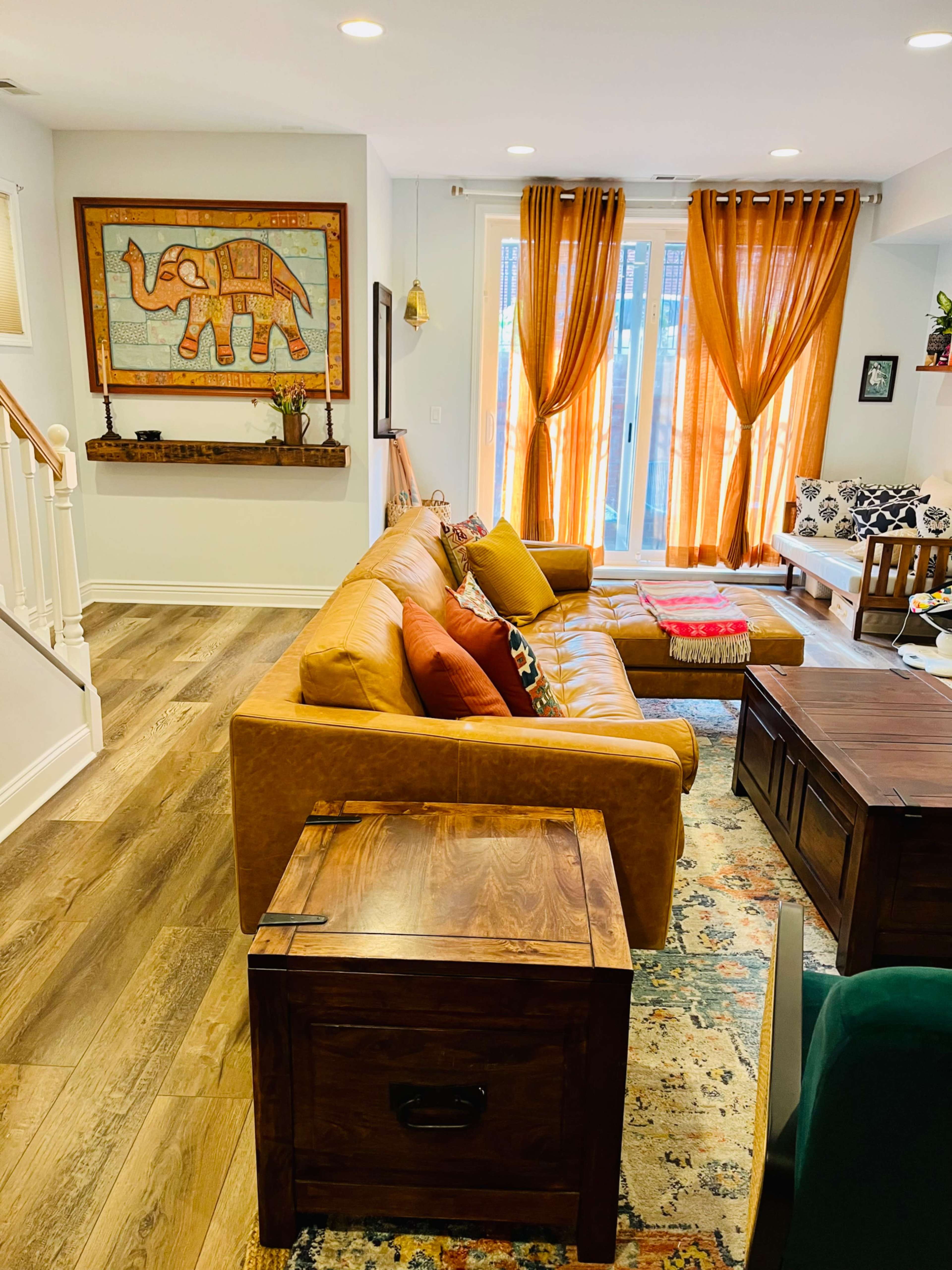 A warmly decorated living room with a brown leather sofa, wooden furniture, and large windows draped with orange curtains.