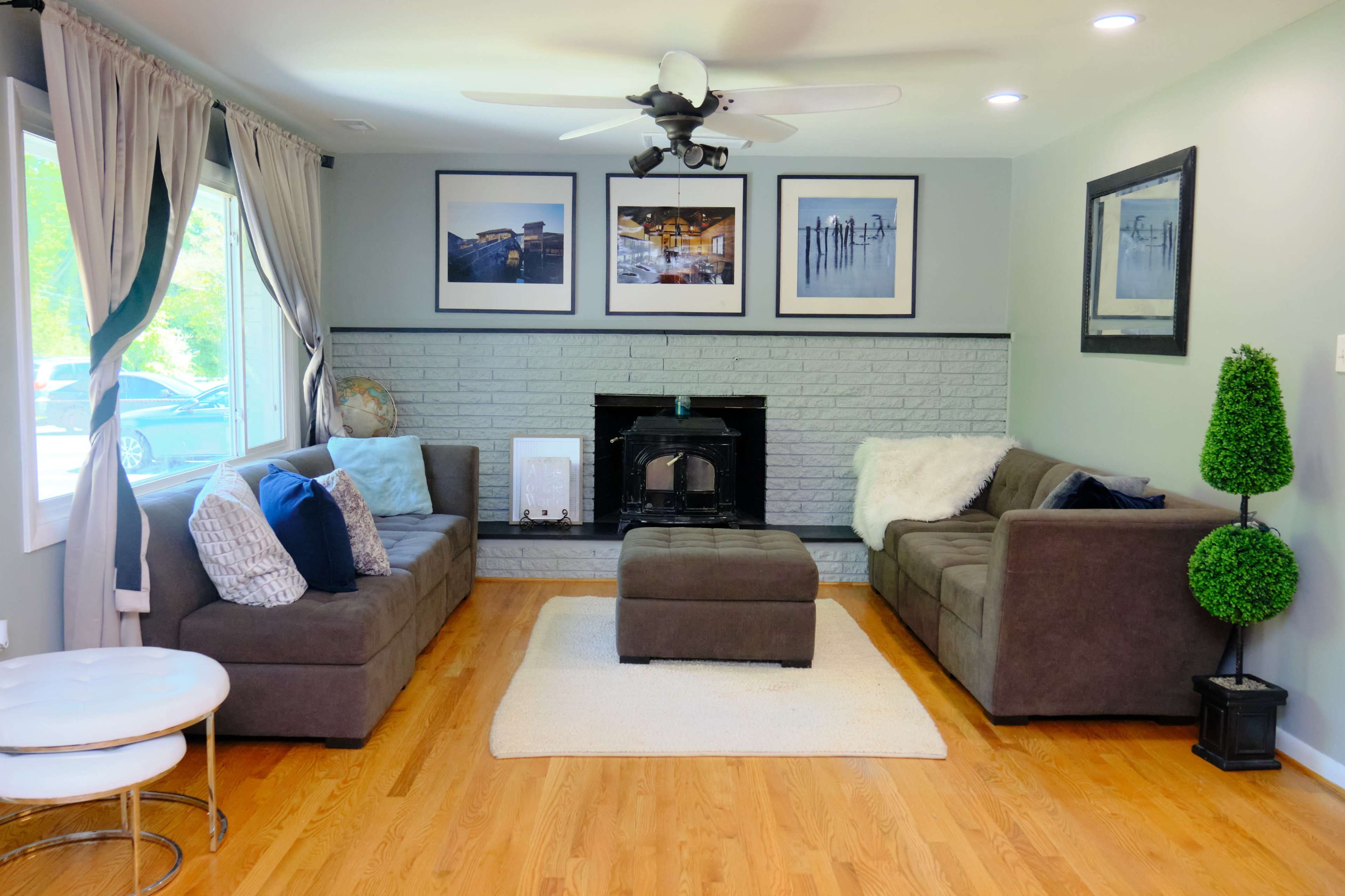 The living room features two dark sofas arranged around a central ottoman, with a wall-mounted fireplace and framed pictures above it.