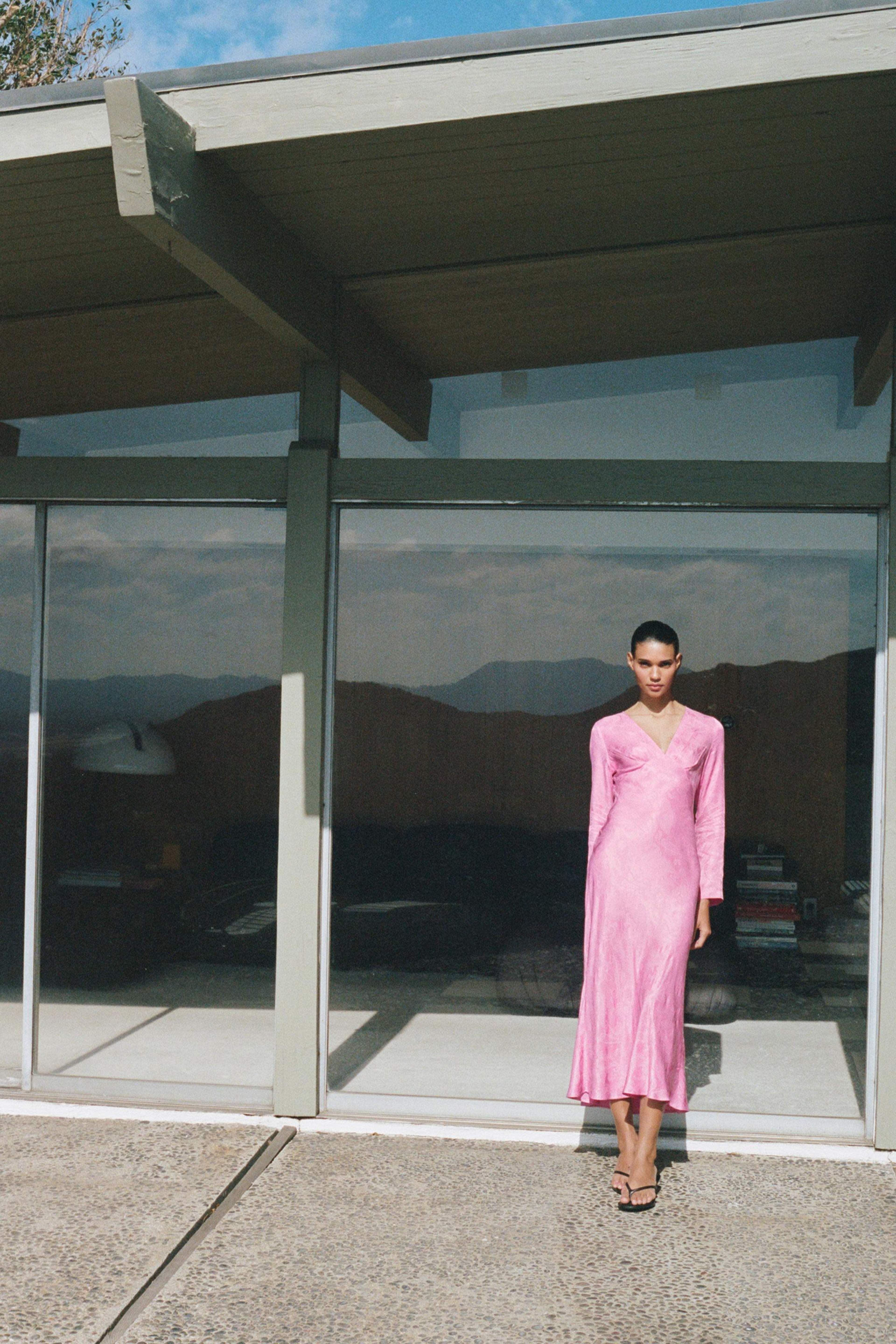 A person in a long pink dress stands in front of a modern glass structure.