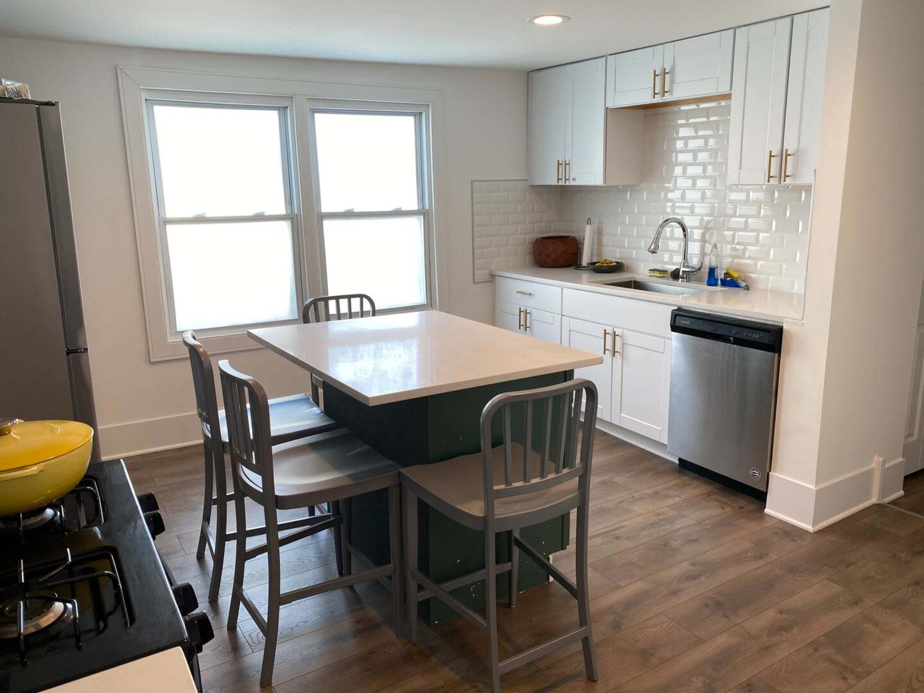 A modern kitchen features white cabinetry, a central island with seating, and large windows allowing natural light to enter.