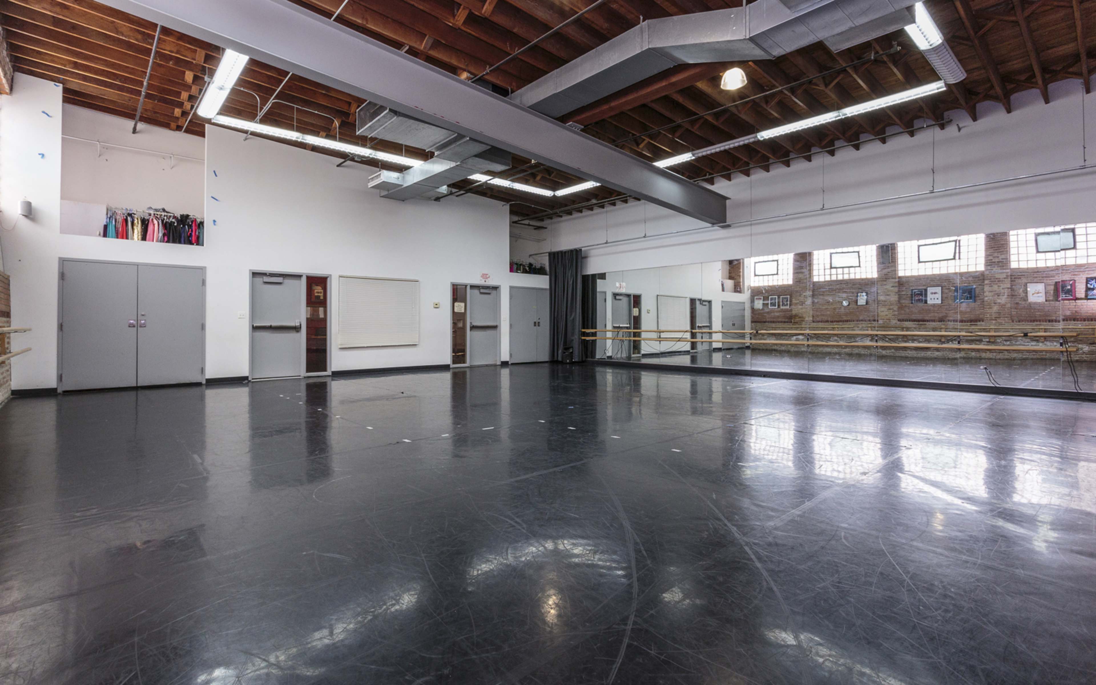 A spacious dance studio with a black vinyl floor, mirrored walls, and large windows allowing natural light to enter.