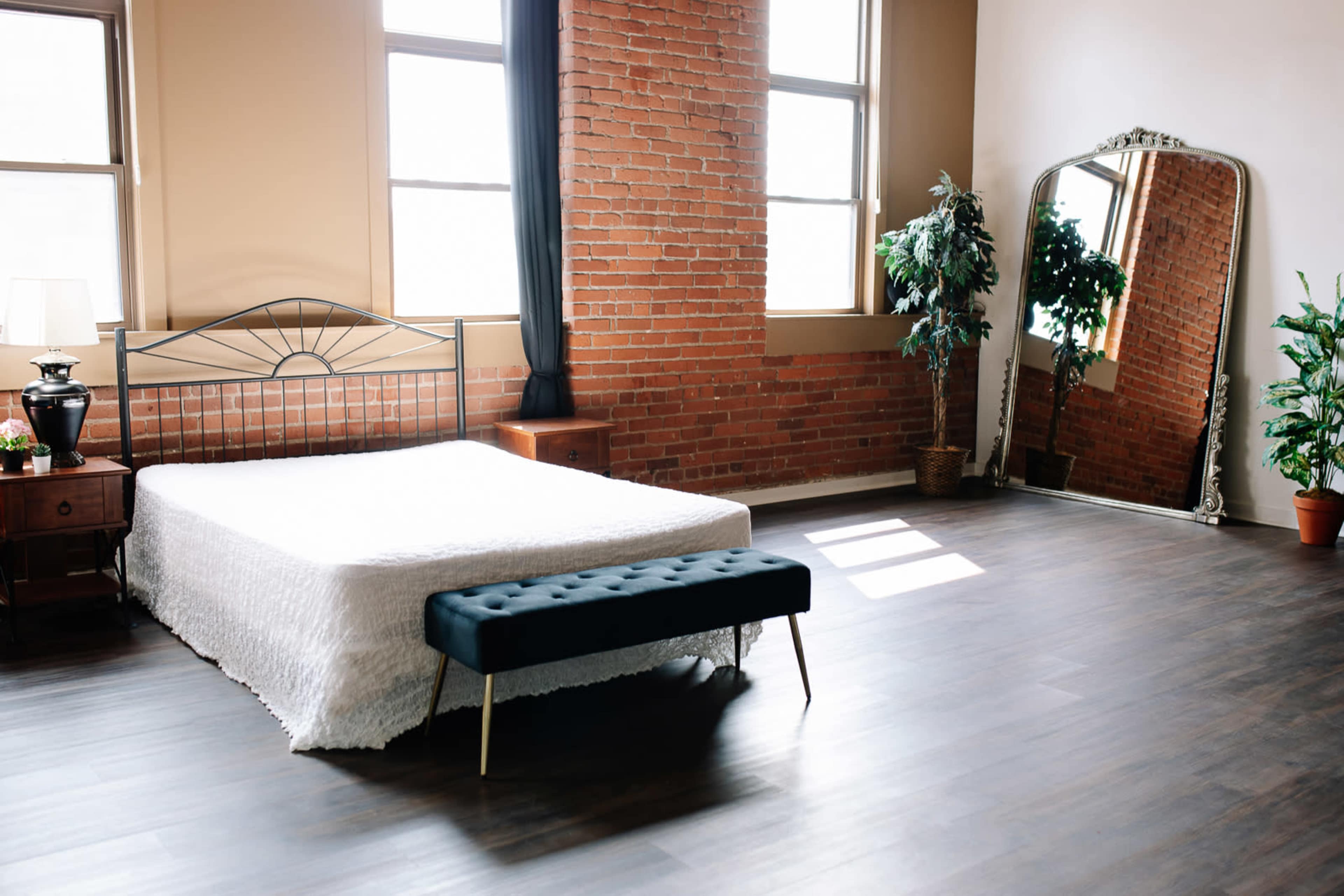 A minimalist bedroom features a metal bed frame with a white bedspread, a tufted black bench, and large windows alongside exposed brick walls.