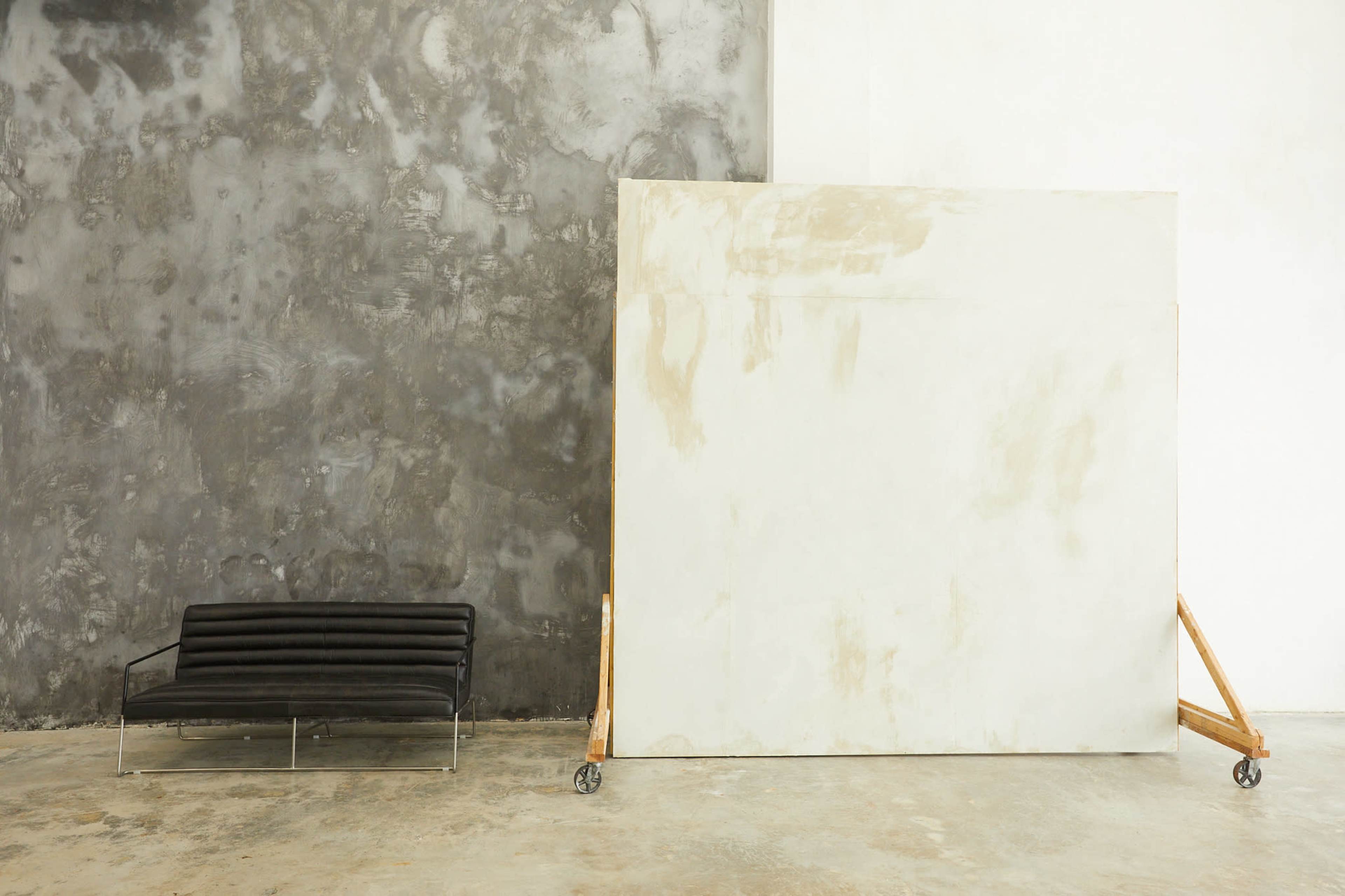 A black sofa is positioned next to a large, unpainted canvas on wheels against a textured gray wall.