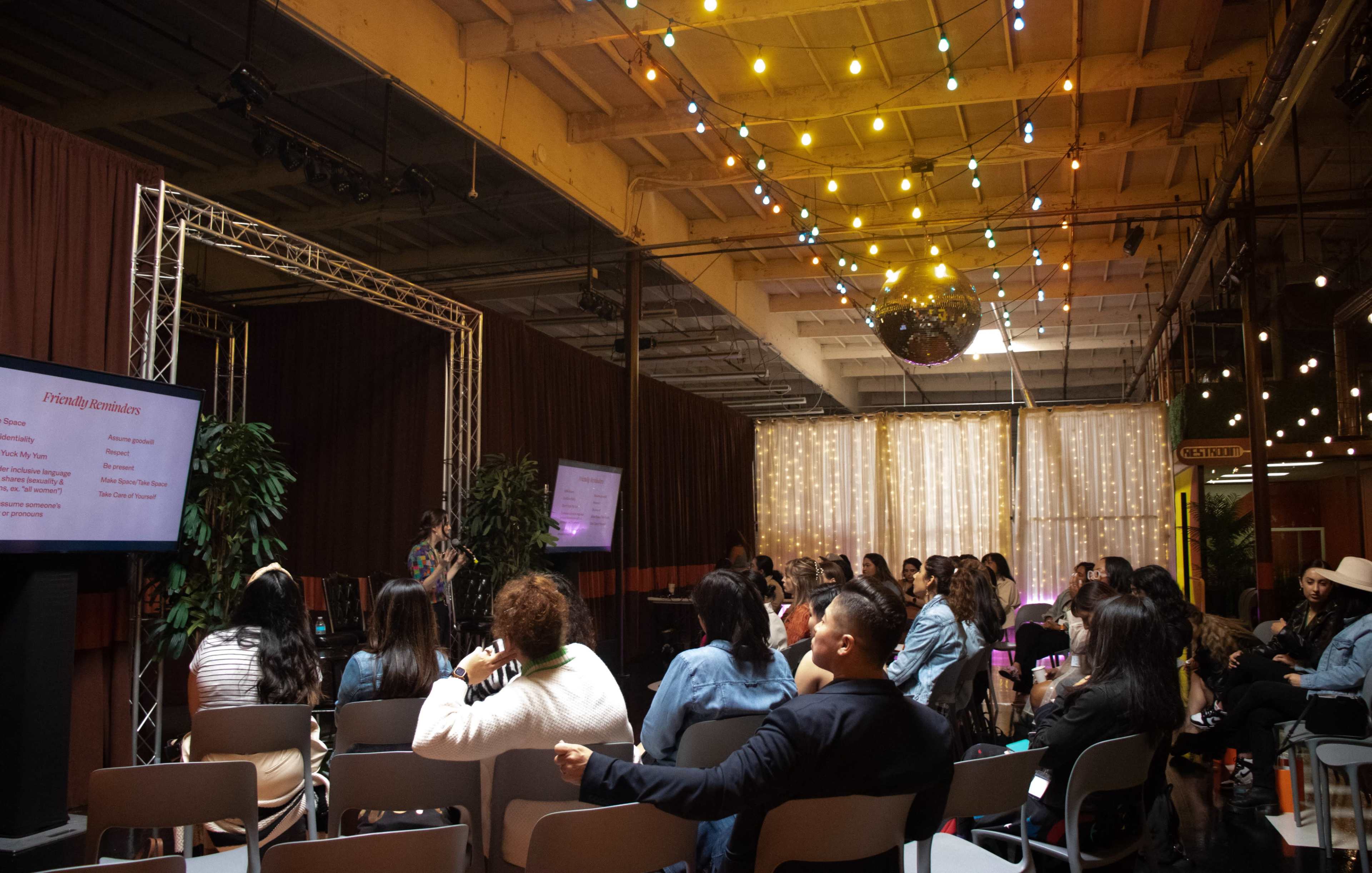 The image shows a group of people seated in a dimly lit venue with string lights above, watching a presentation on stage.