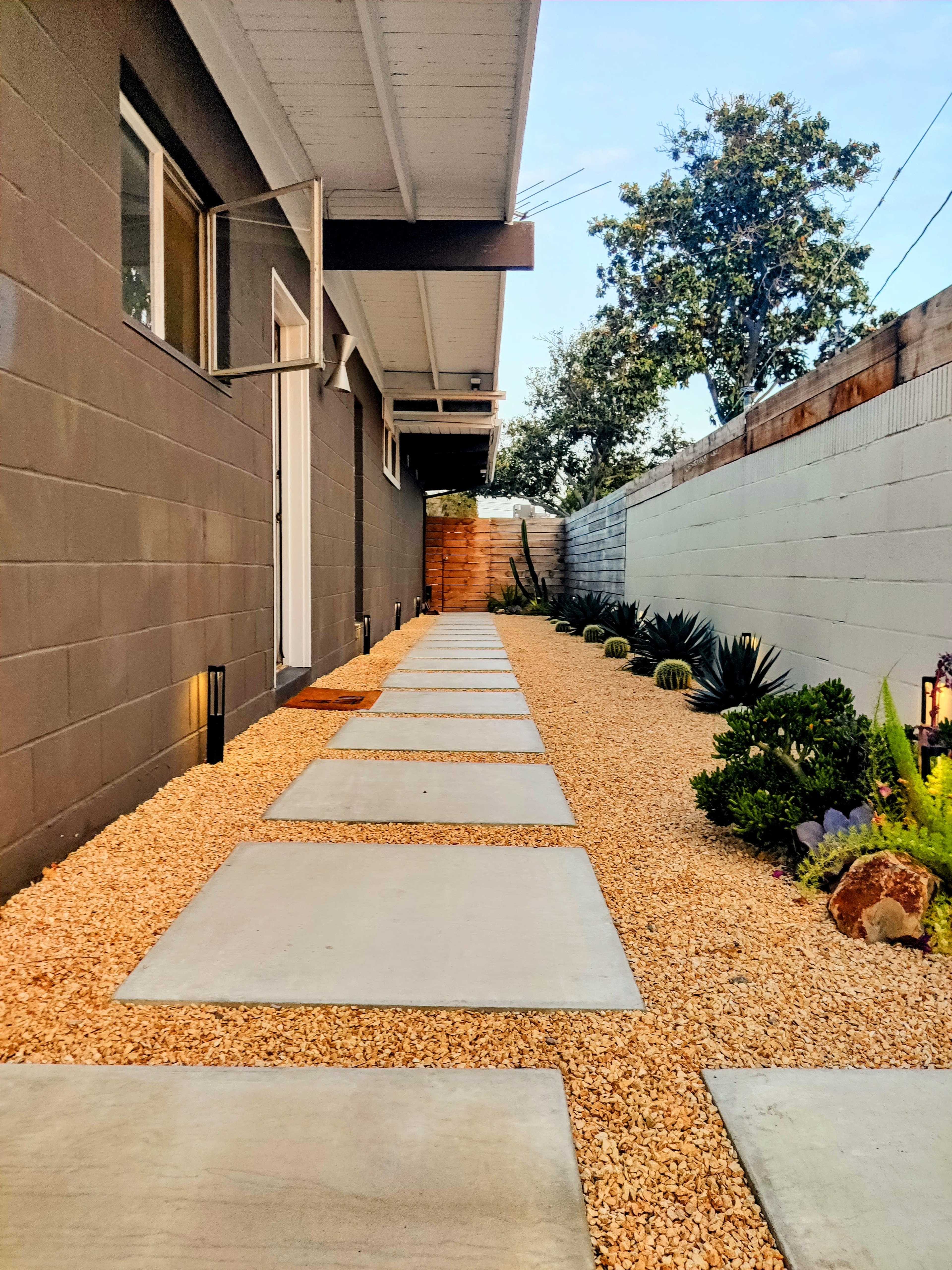The image shows a narrow walkway lined with concrete stepping stones, bordered by gravel and landscaped with plants along a residential building.