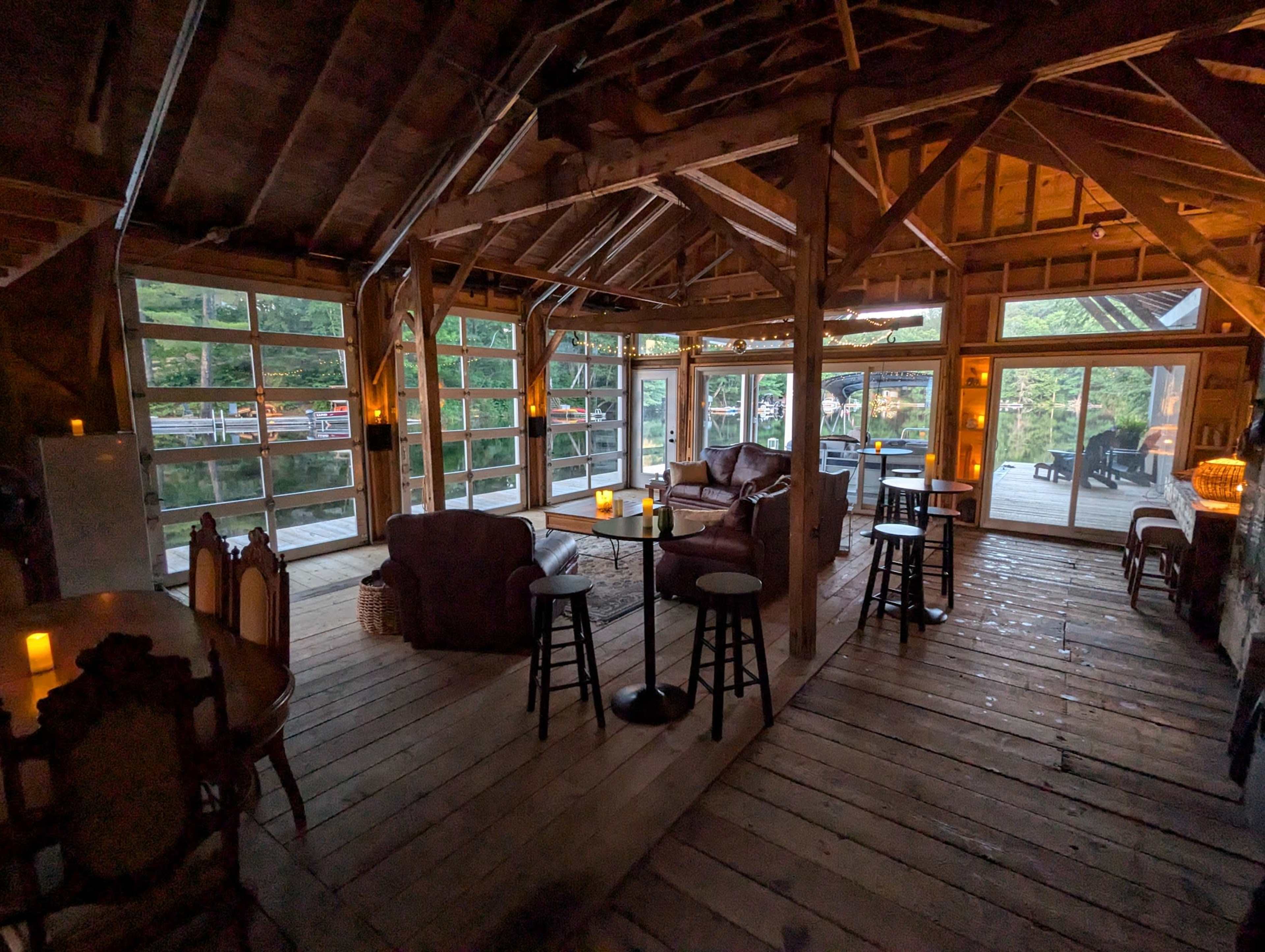 The image shows a rustic interior of a wooden cabin with lounge seating, large windows, and dim lighting.