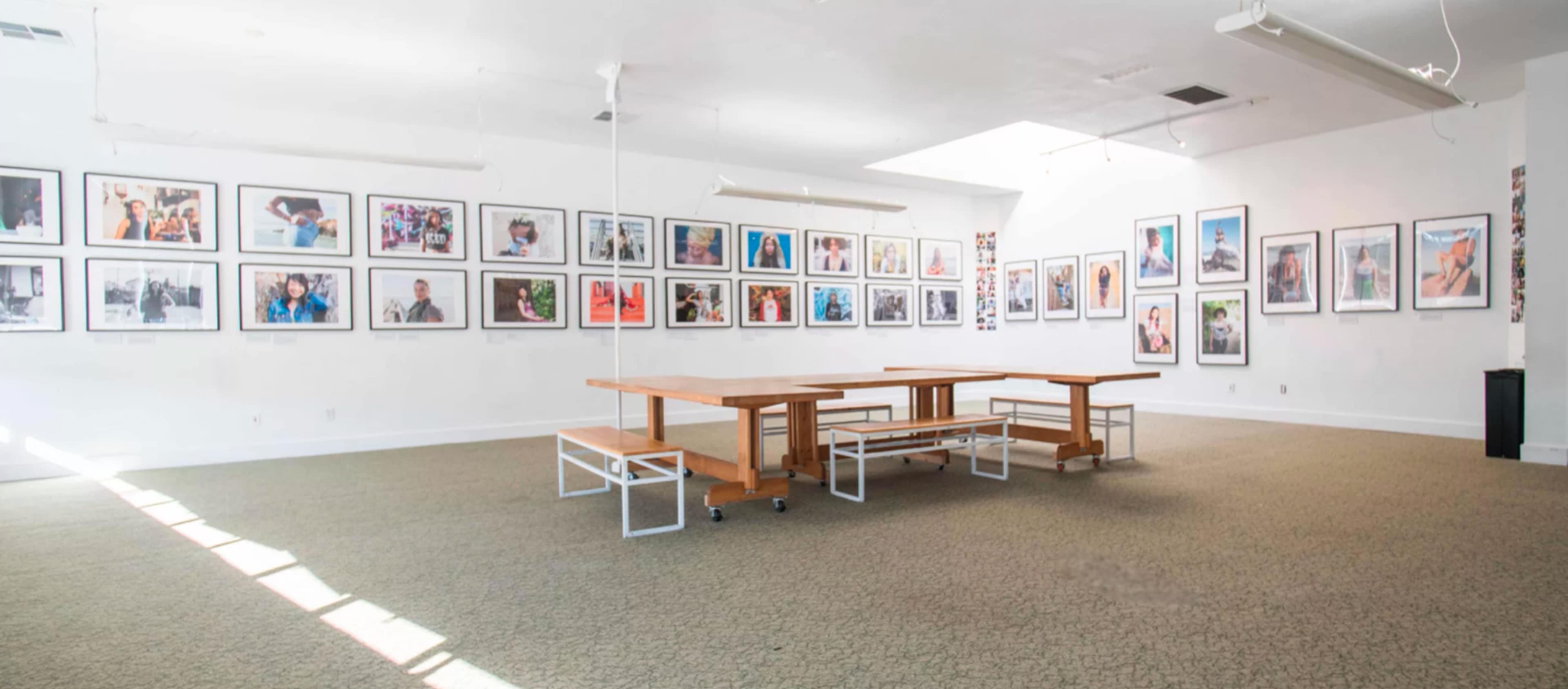 A spacious gallery with several framed photographs displayed on the walls and a large wooden table surrounded by benches in the center.