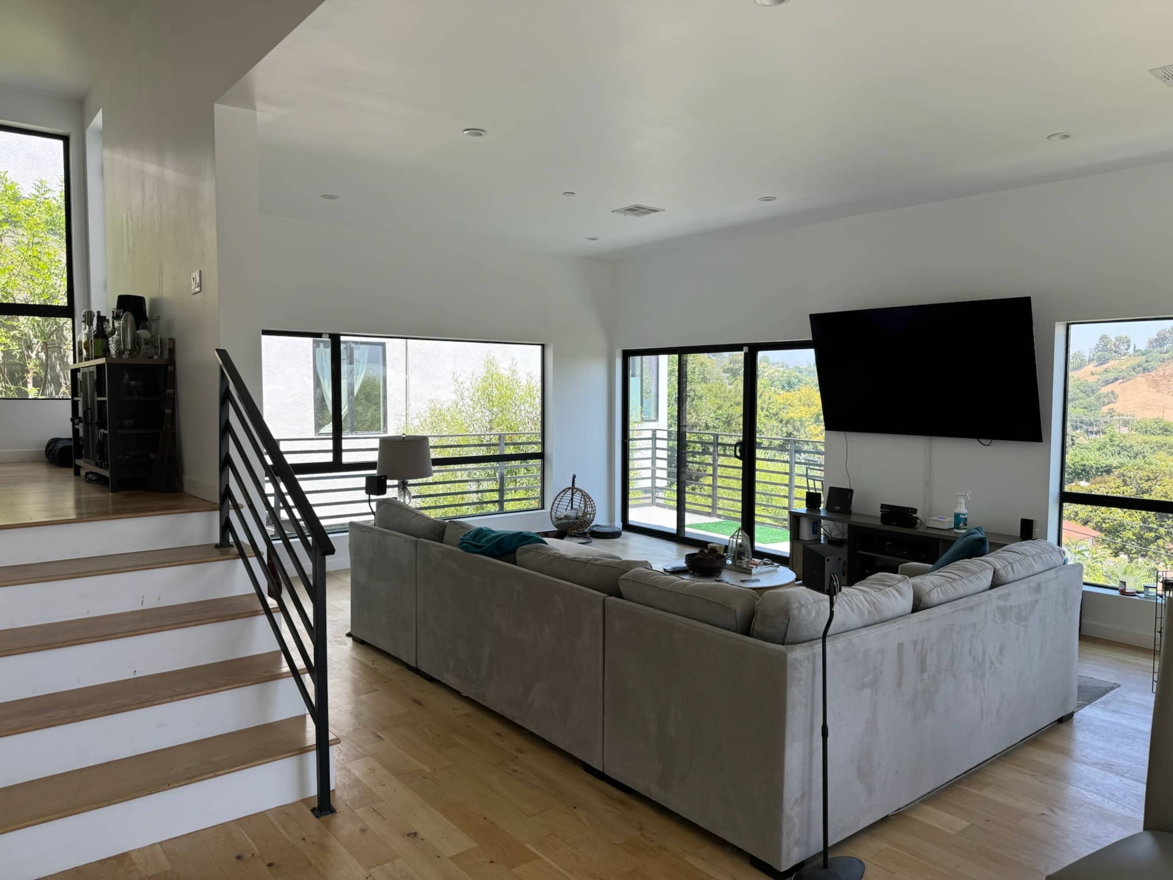The image shows a modern living room with large windows, a sectional sofa, and a staircase leading to an upper level.