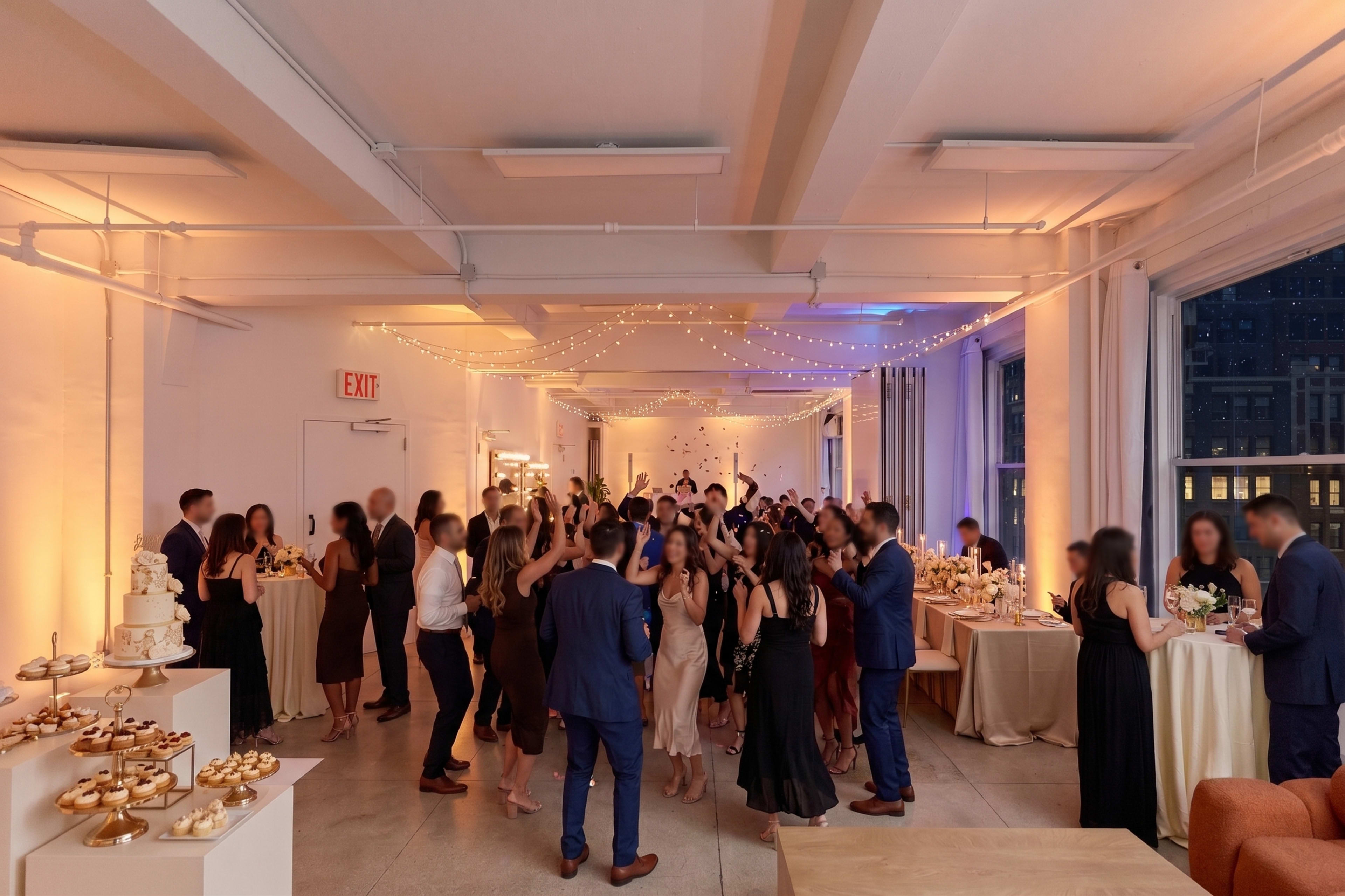 A large gathering of people is dancing and socializing in a well-lit event space adorned with decorative lighting and tables of food.