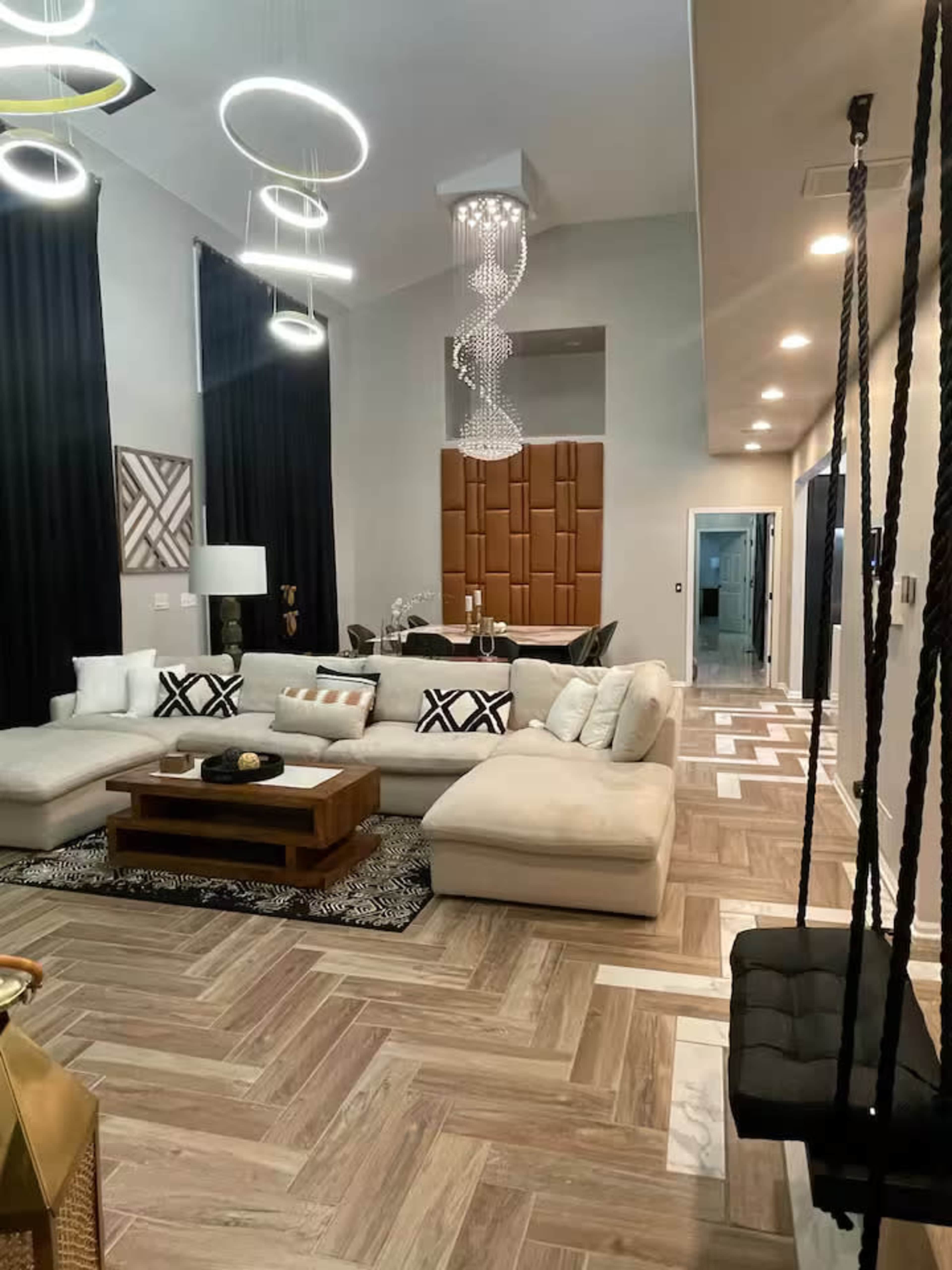 The image shows a modern living room featuring a large sectional sofa, a coffee table, decorative pillows, and stylish lighting fixtures hanging from the ceiling.