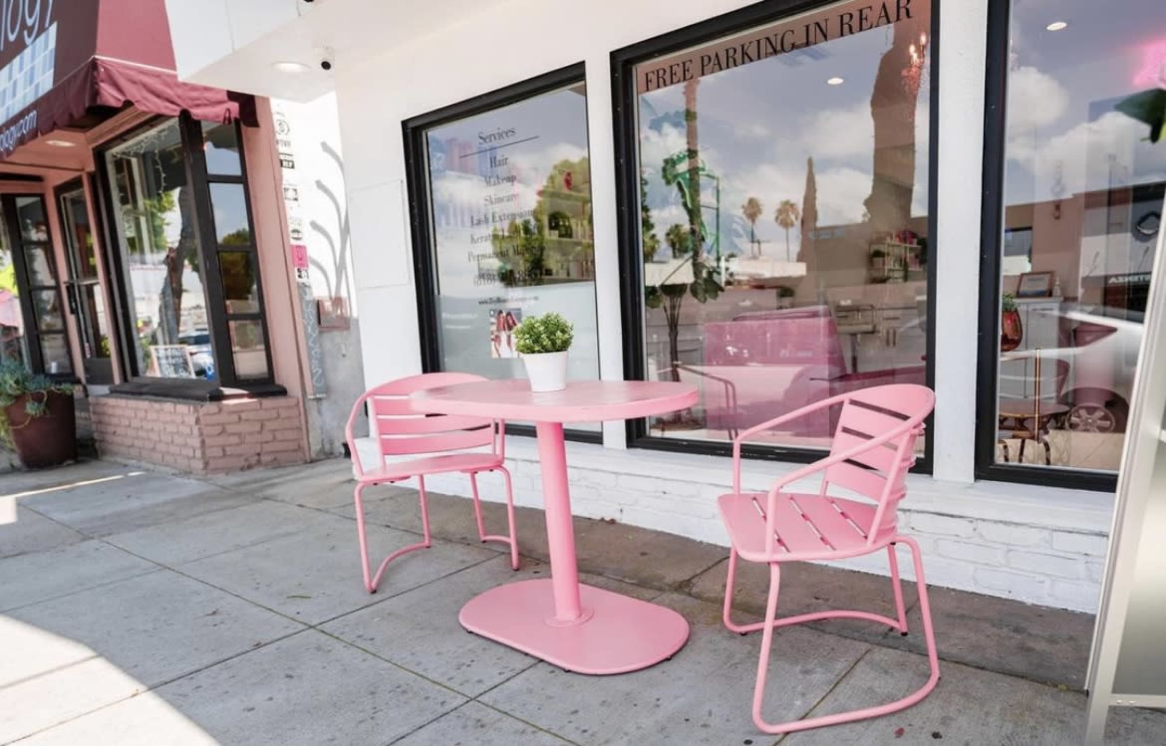 A small pink outdoor table with two matching chairs sits on a sidewalk in front of a store, accompanied by a potted plant.
