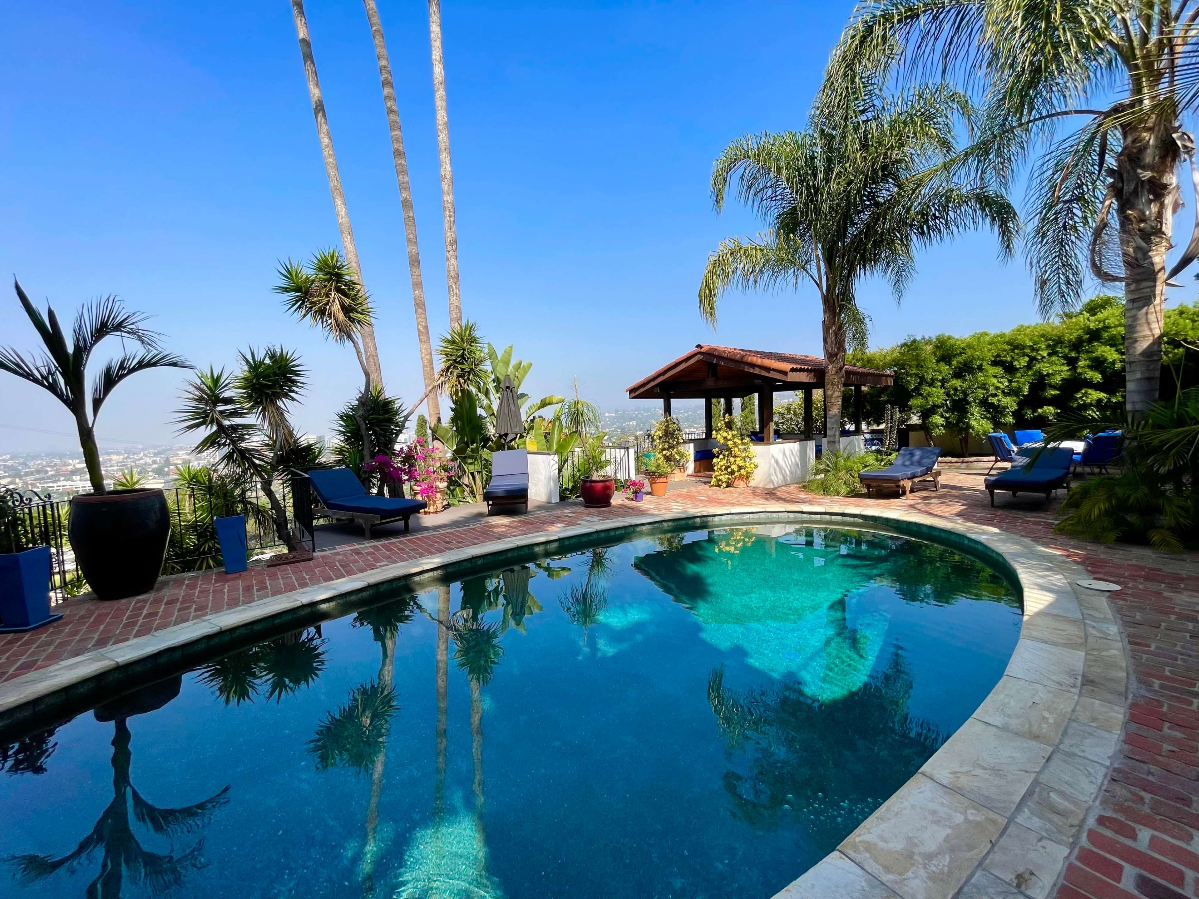 The image shows a luxurious swimming pool surrounded by palm trees and tropical plants, with a shaded patio area in the background.