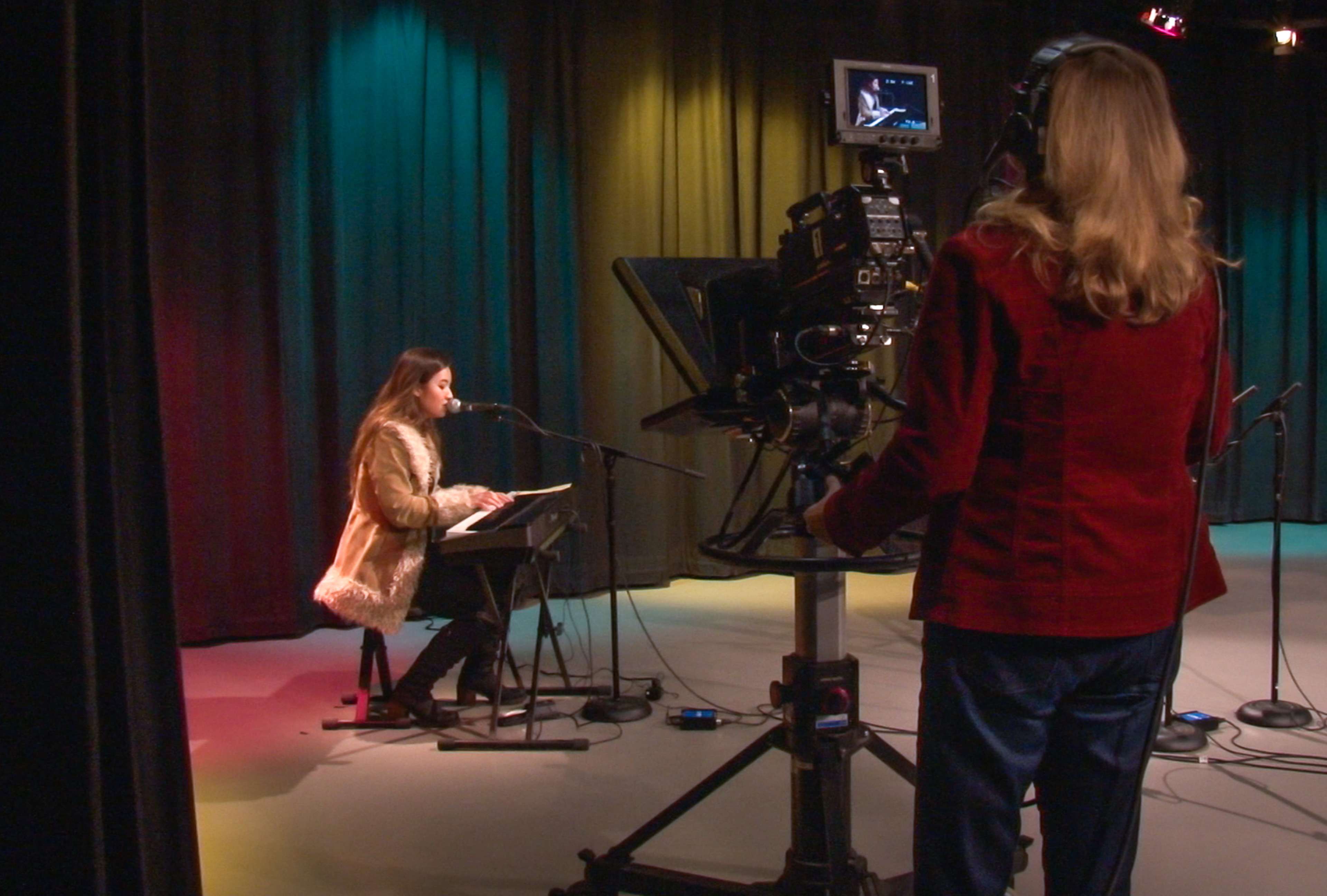 A woman in a faux fur coat performs at a microphone while another woman operates a video camera in a studio with colored backdrop lights.
