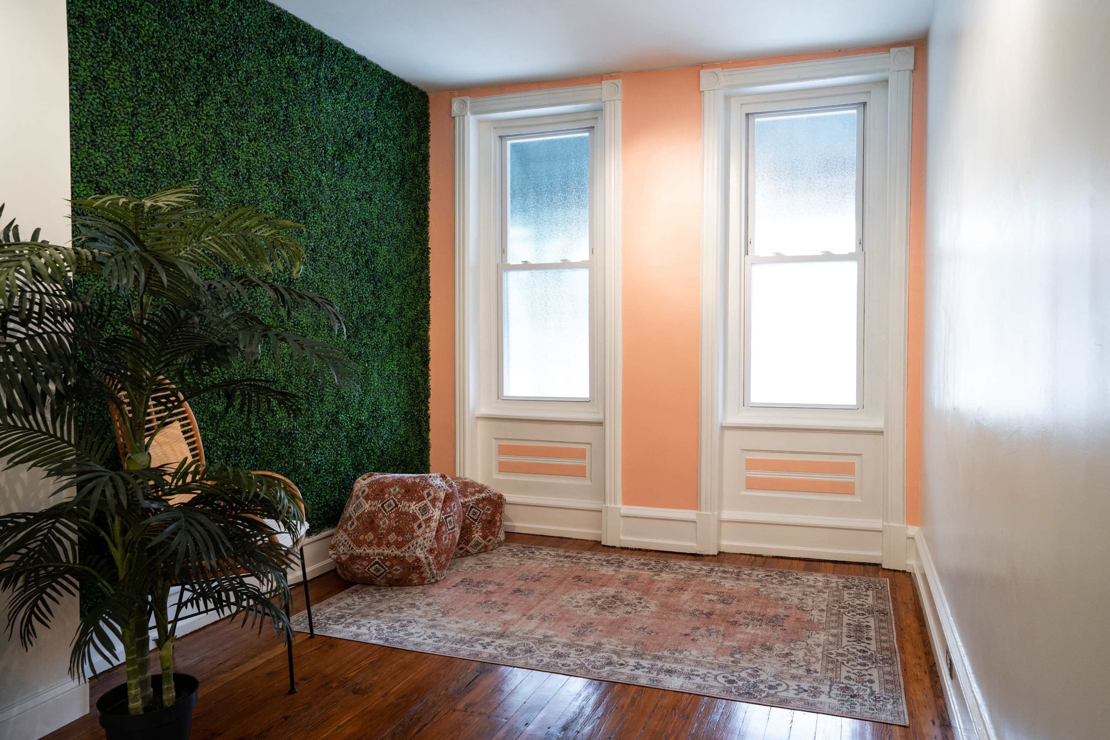 A corner of a room featuring a green plant wall, two windows with white frames, and an orange-painted wall with a patterned rug on the wooden floor.