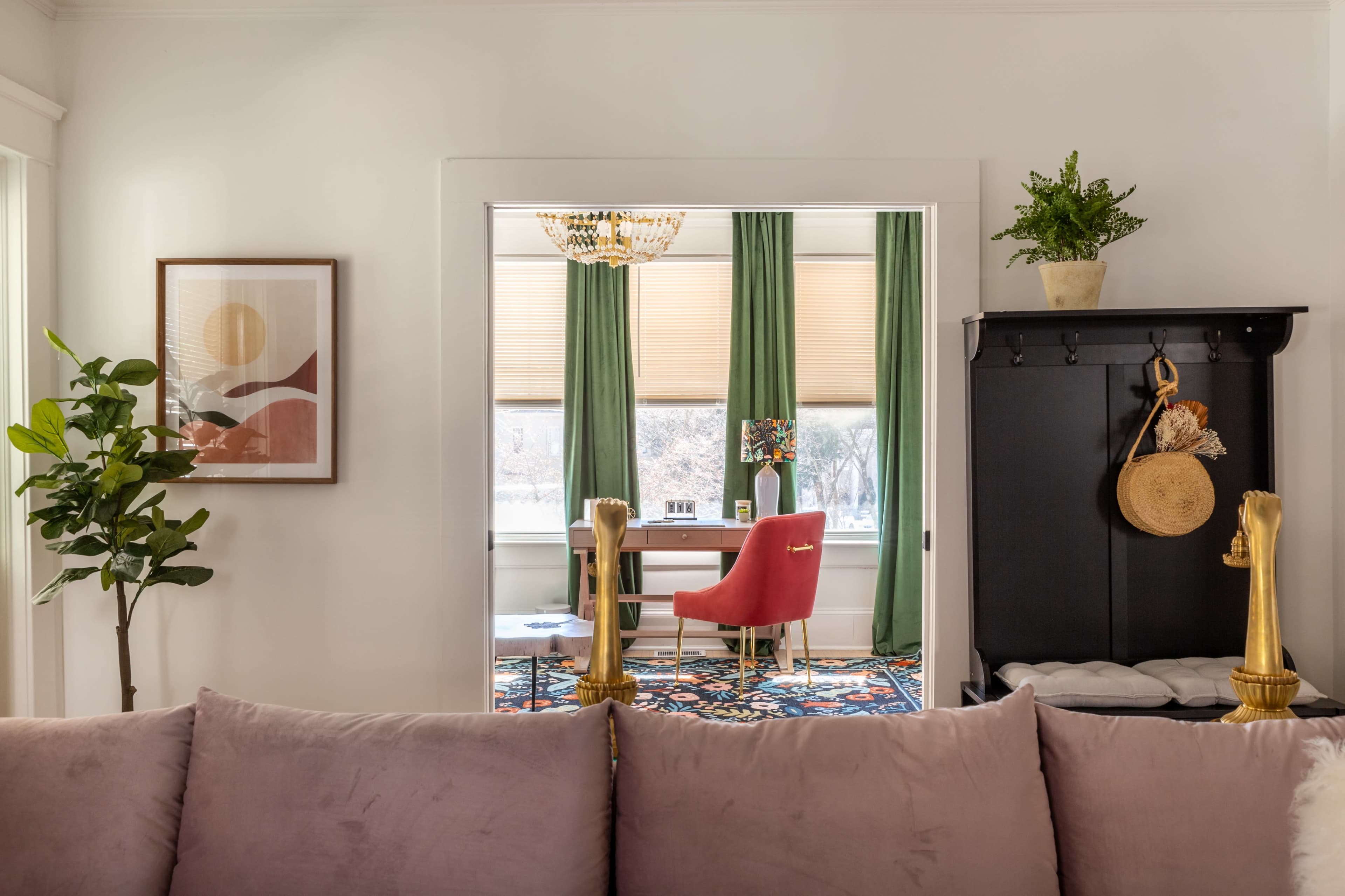 A living room with a view of a home office, featuring a red chair and a desk through an open doorway, while a plant and framed artwork decorate the foreground.