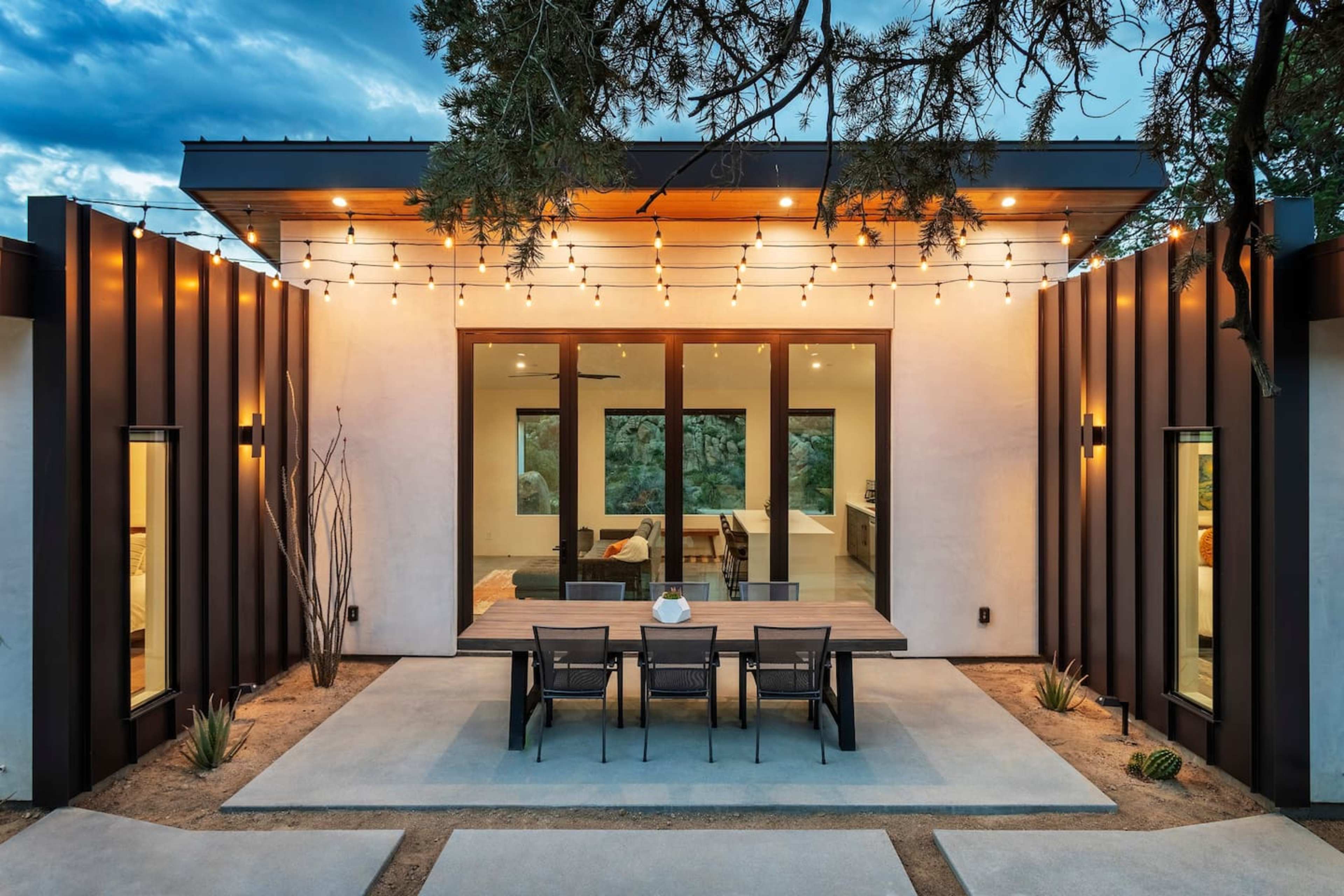 A modern home with a patio area featuring a large table and string lights, surrounded by desert landscaping.