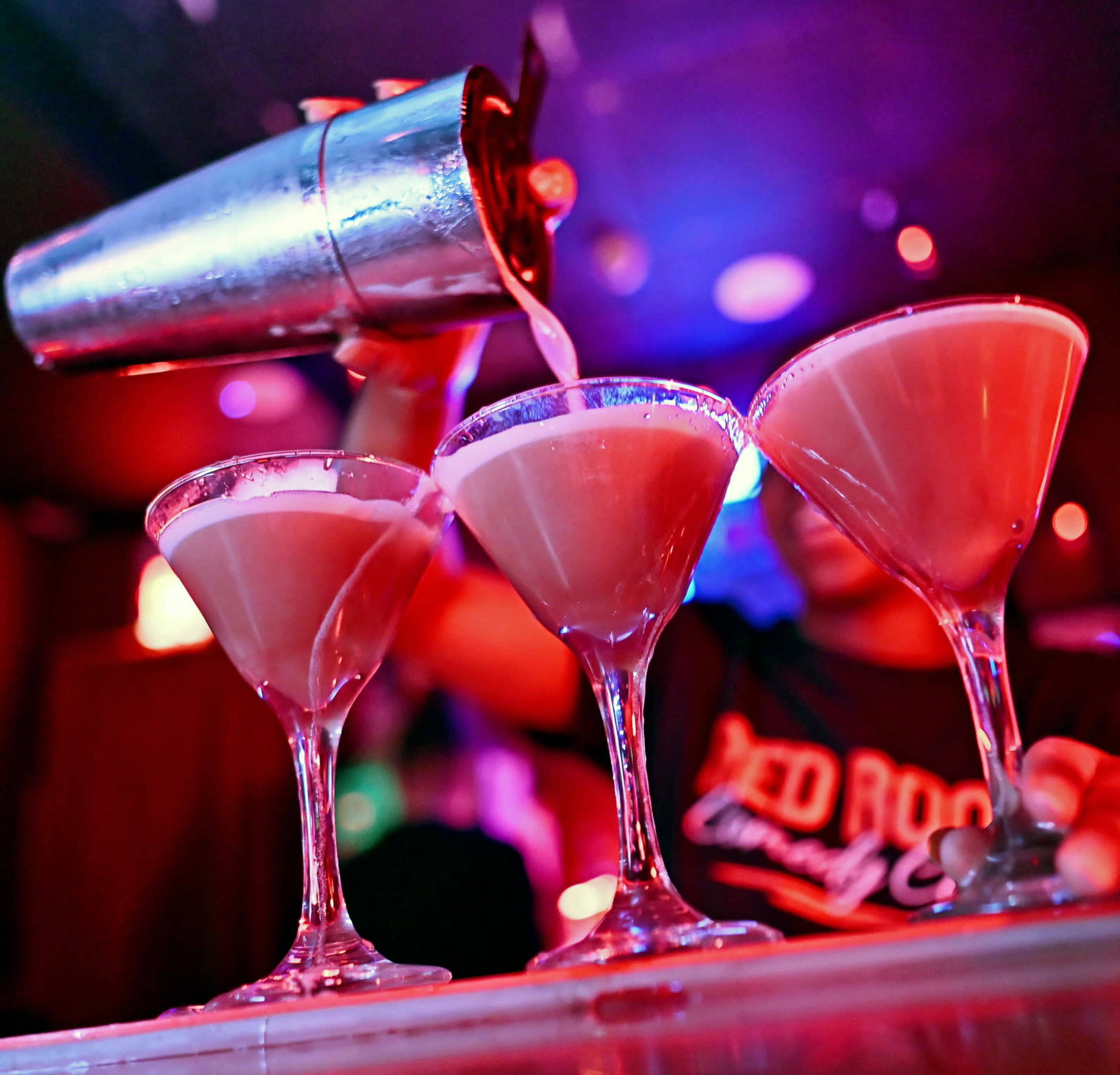A bartender pours a pink cocktail from a shaker into three martini glasses in a brightly lit bar.
