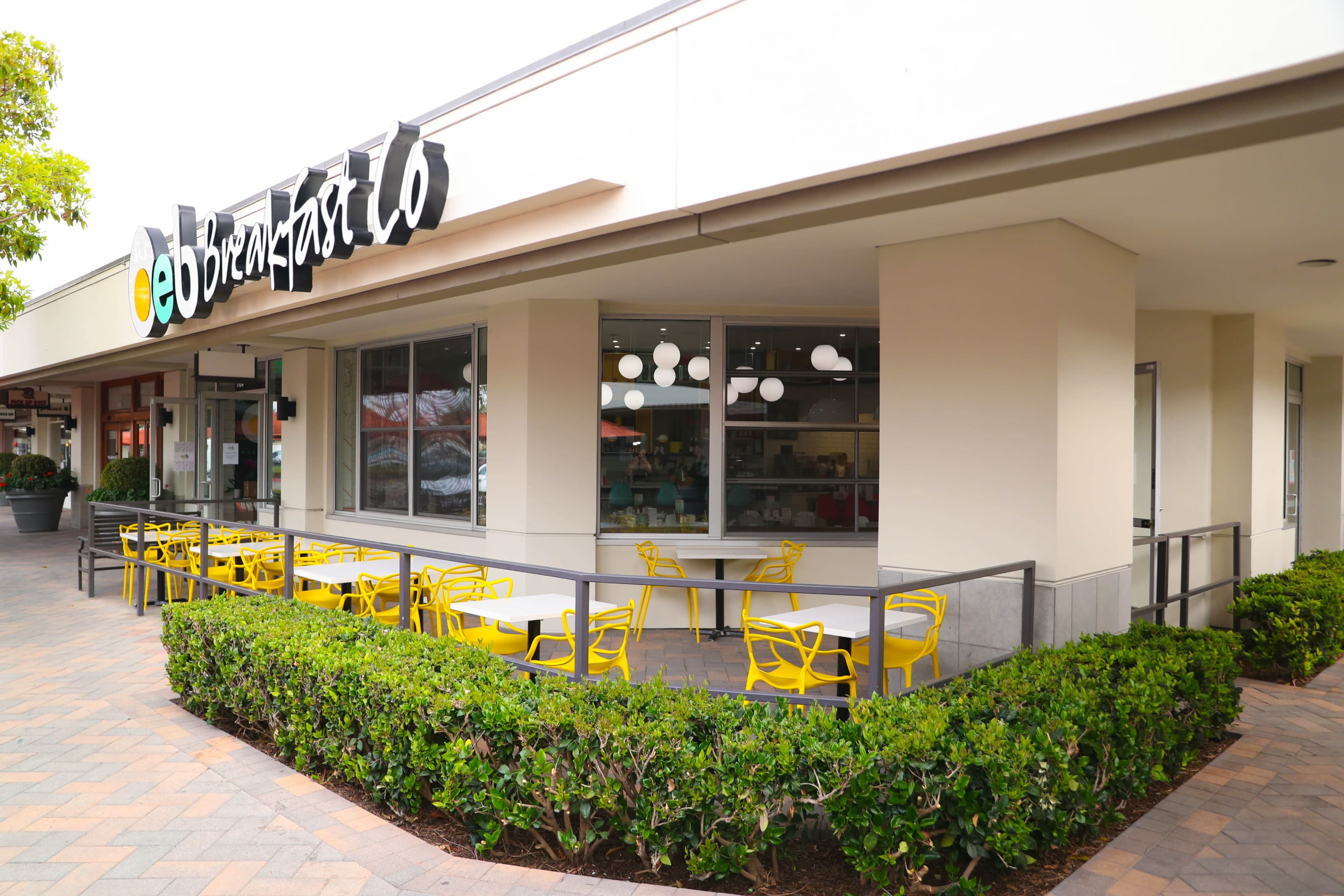 A modern restaurant exterior with yellow chairs and tables arranged outside, surrounded by a neatly trimmed hedge.