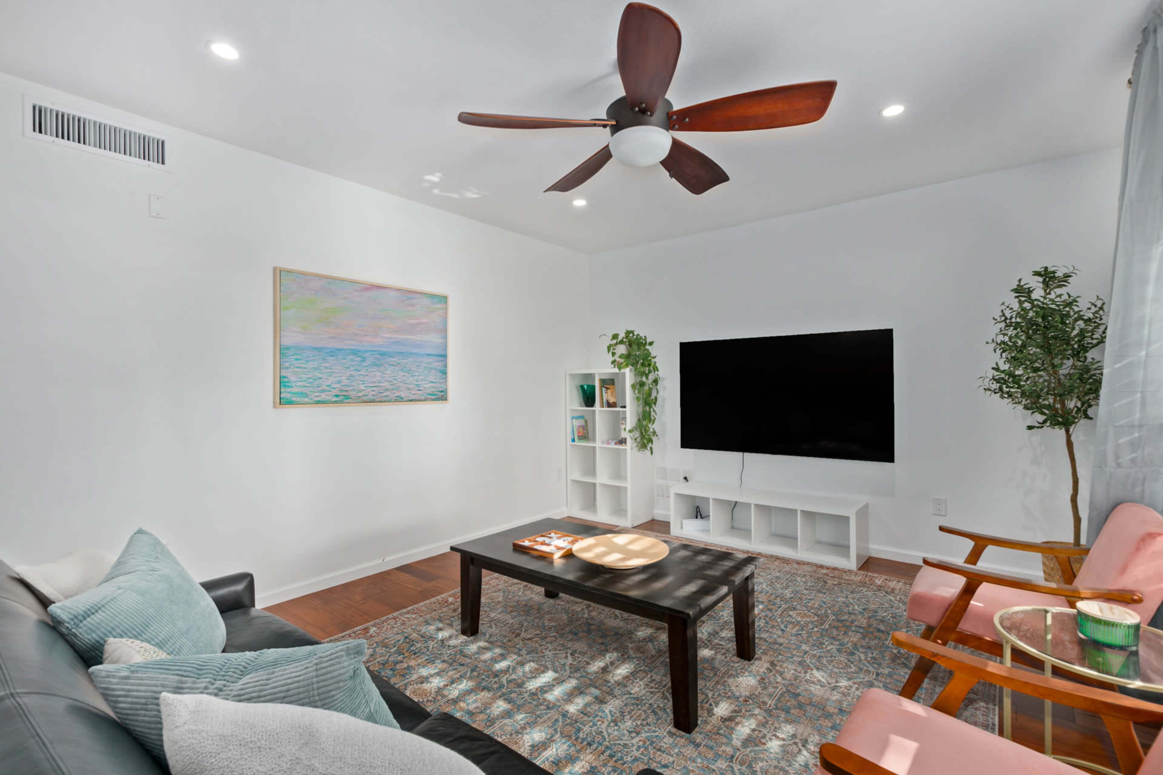 The room features a cozy living area with a black sofa, two wooden chairs, a large TV mounted on the wall, and decorative plants.