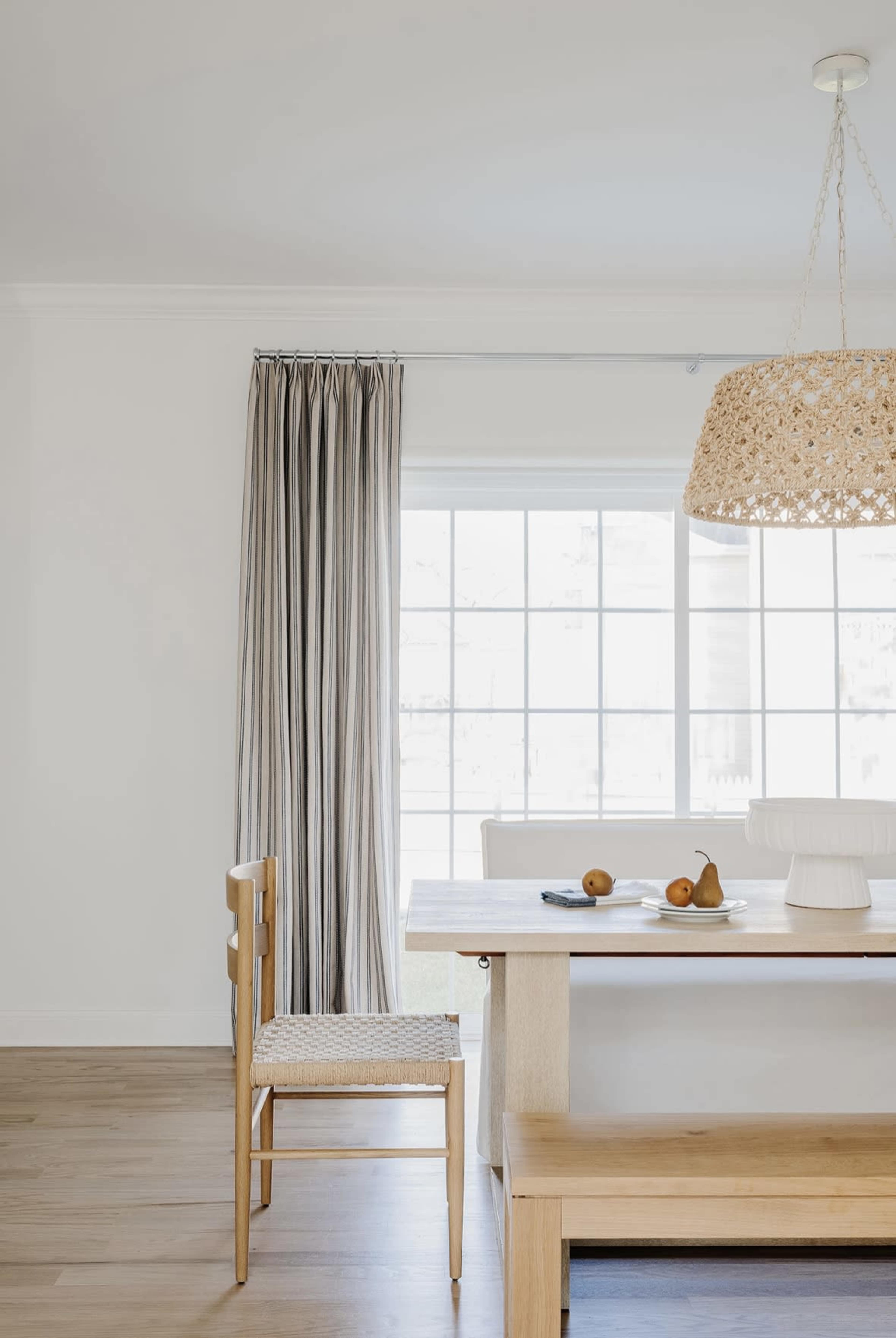 A minimalist dining area features a wooden table, a chair with a woven seat, and a pendant light illuminating the space.