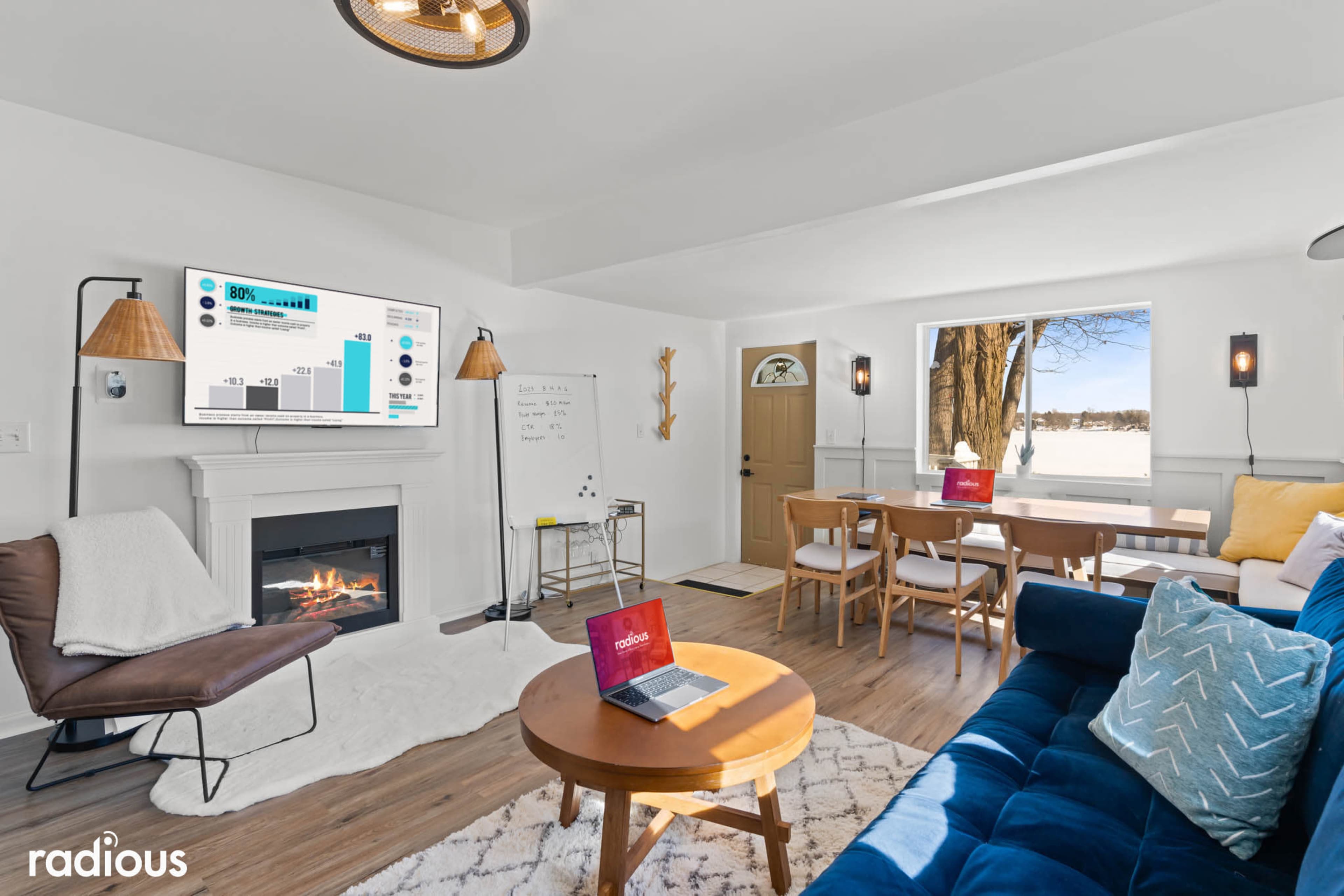 A modern living room with a fireplace, a wall-mounted screen displaying data, a laptop on a table, and a dining area with a wooden table and chairs.