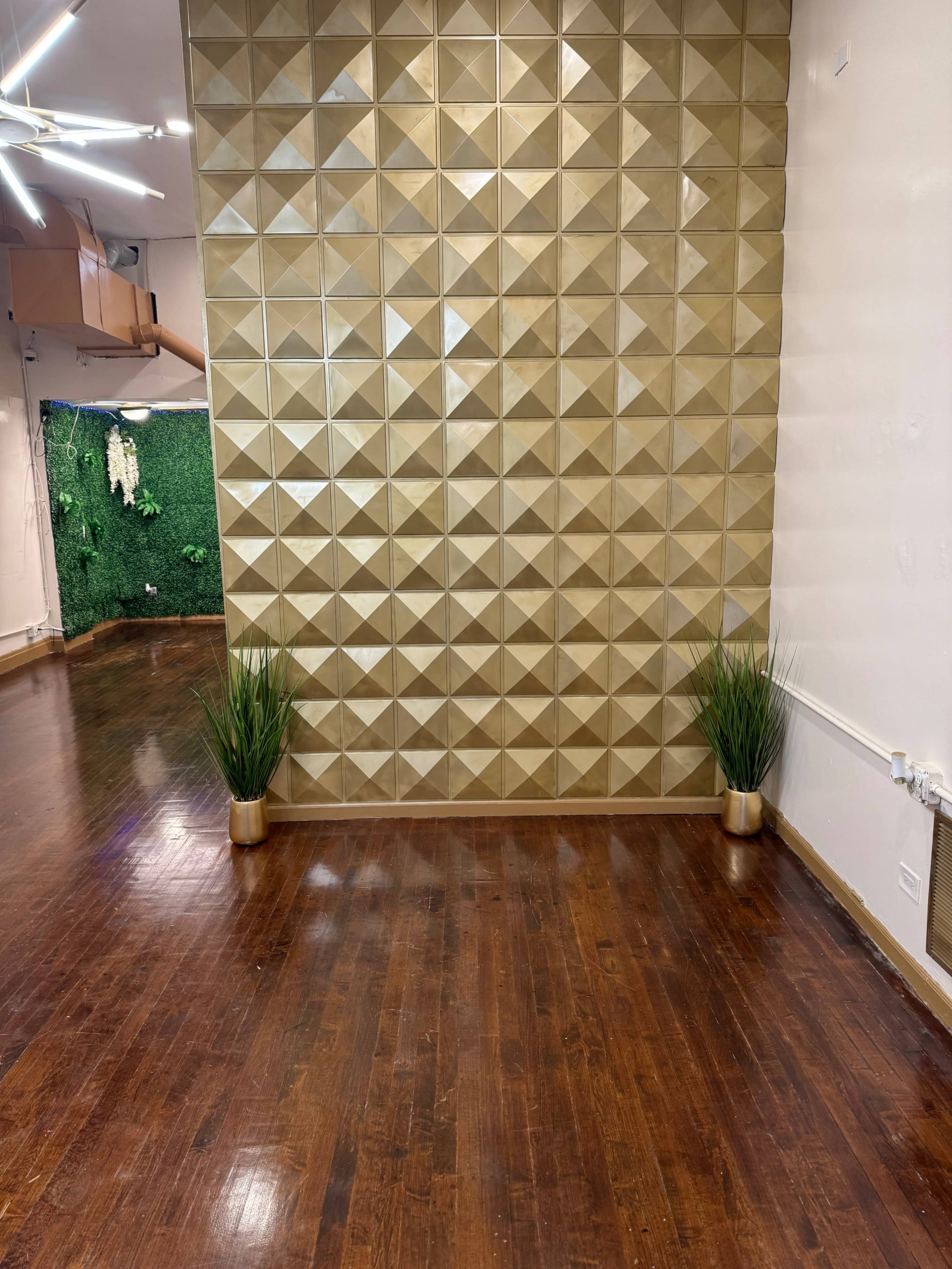 A room with a golden, textured wall on one side, wooden flooring, and two small potted plants placed in the corners.
