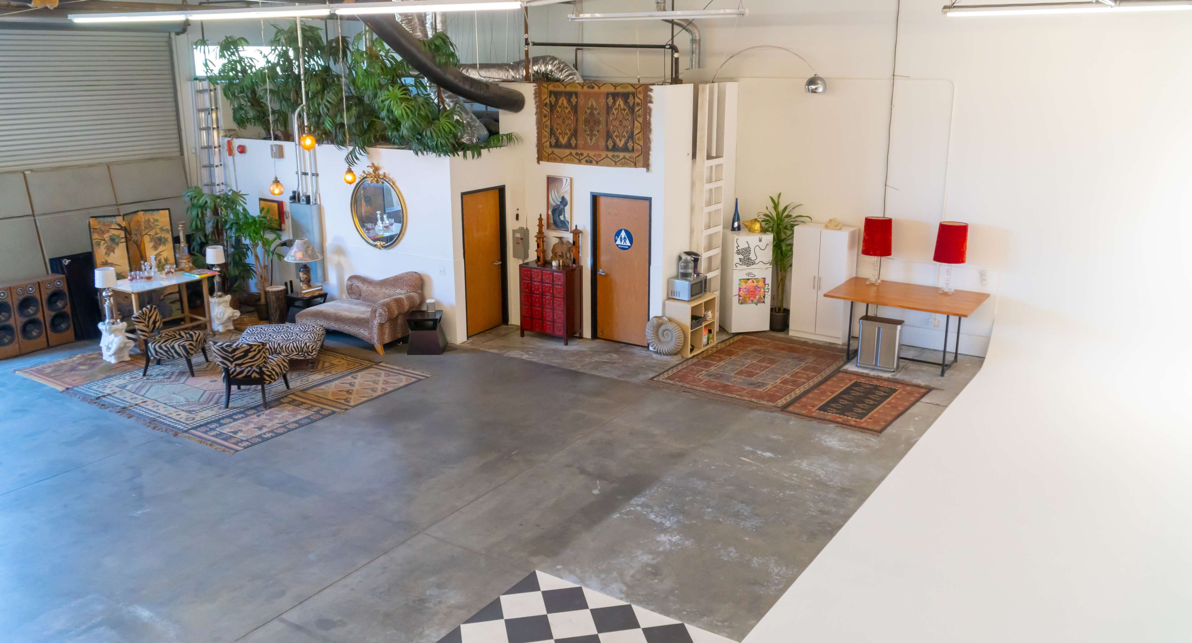 A spacious warehouse interior featuring a seating area with a sofa, decorative rugs, and plants, along with a separate room entrance and tables.