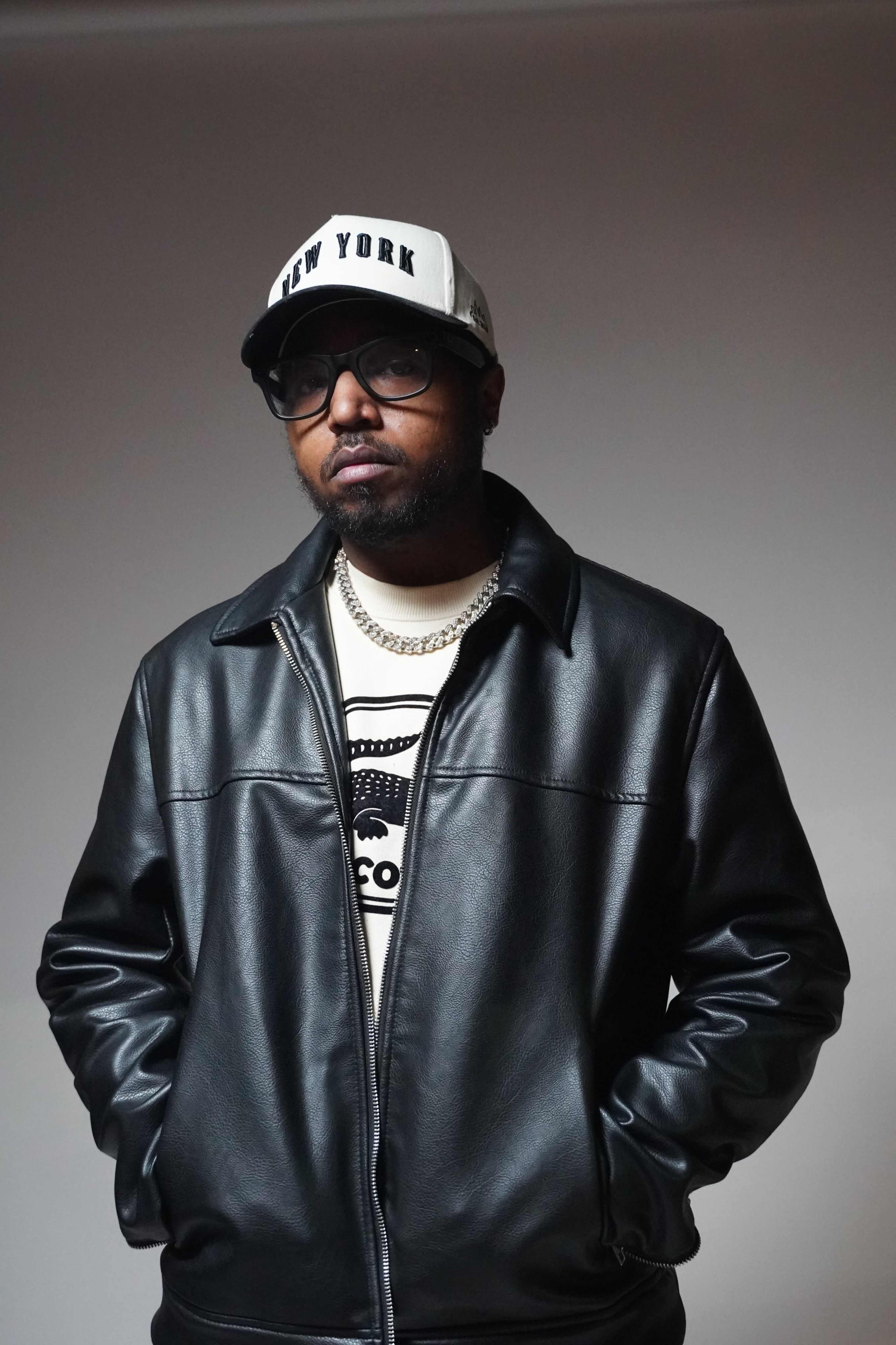 A man stands confidently in a black leather jacket, wearing a white cap with "NEW YORK" printed on it and a light-colored shirt underneath.