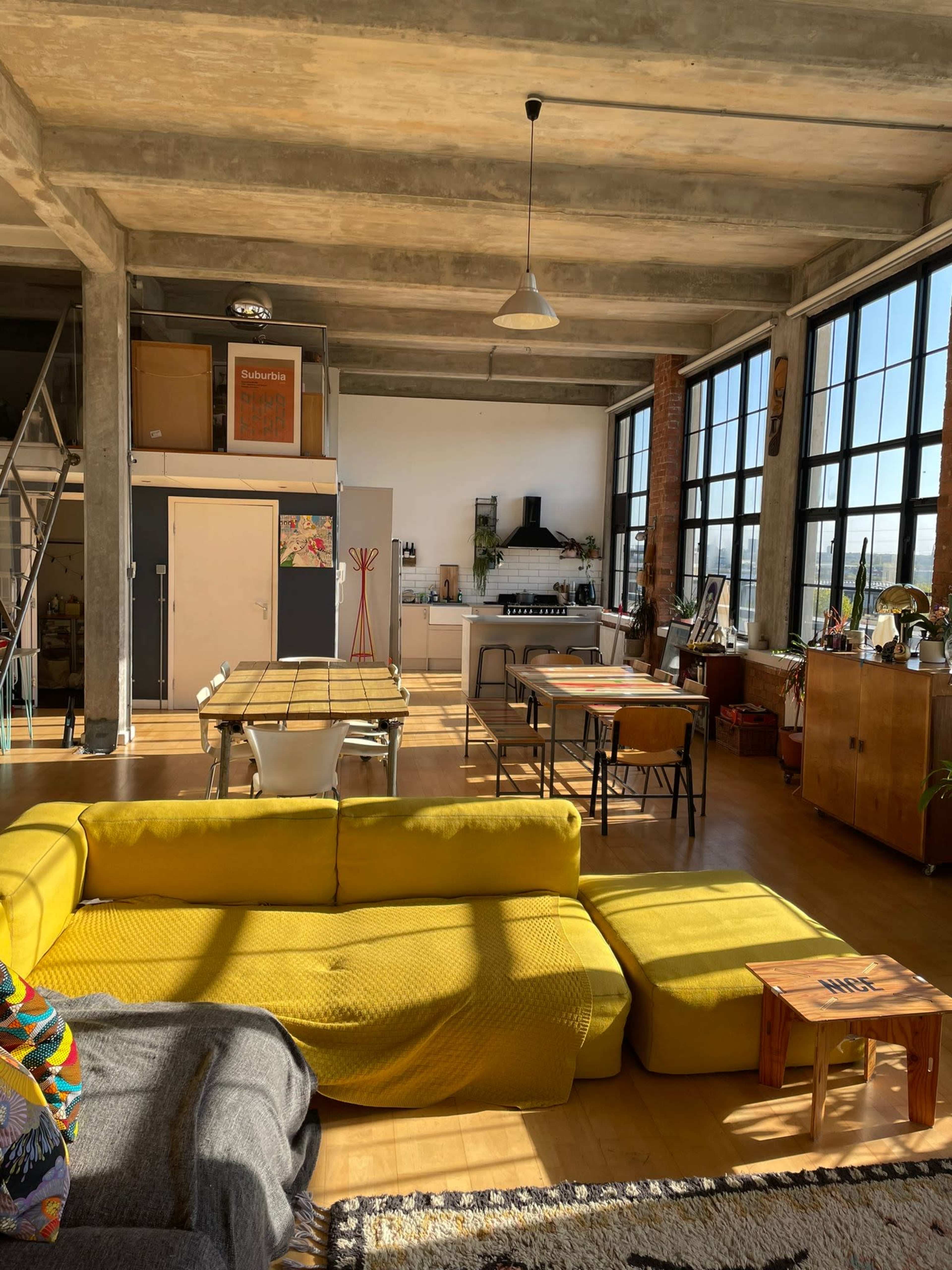 A spacious, modern loft-style living area with large windows, a yellow sectional sofa, a dining table, and a kitchen in the background.