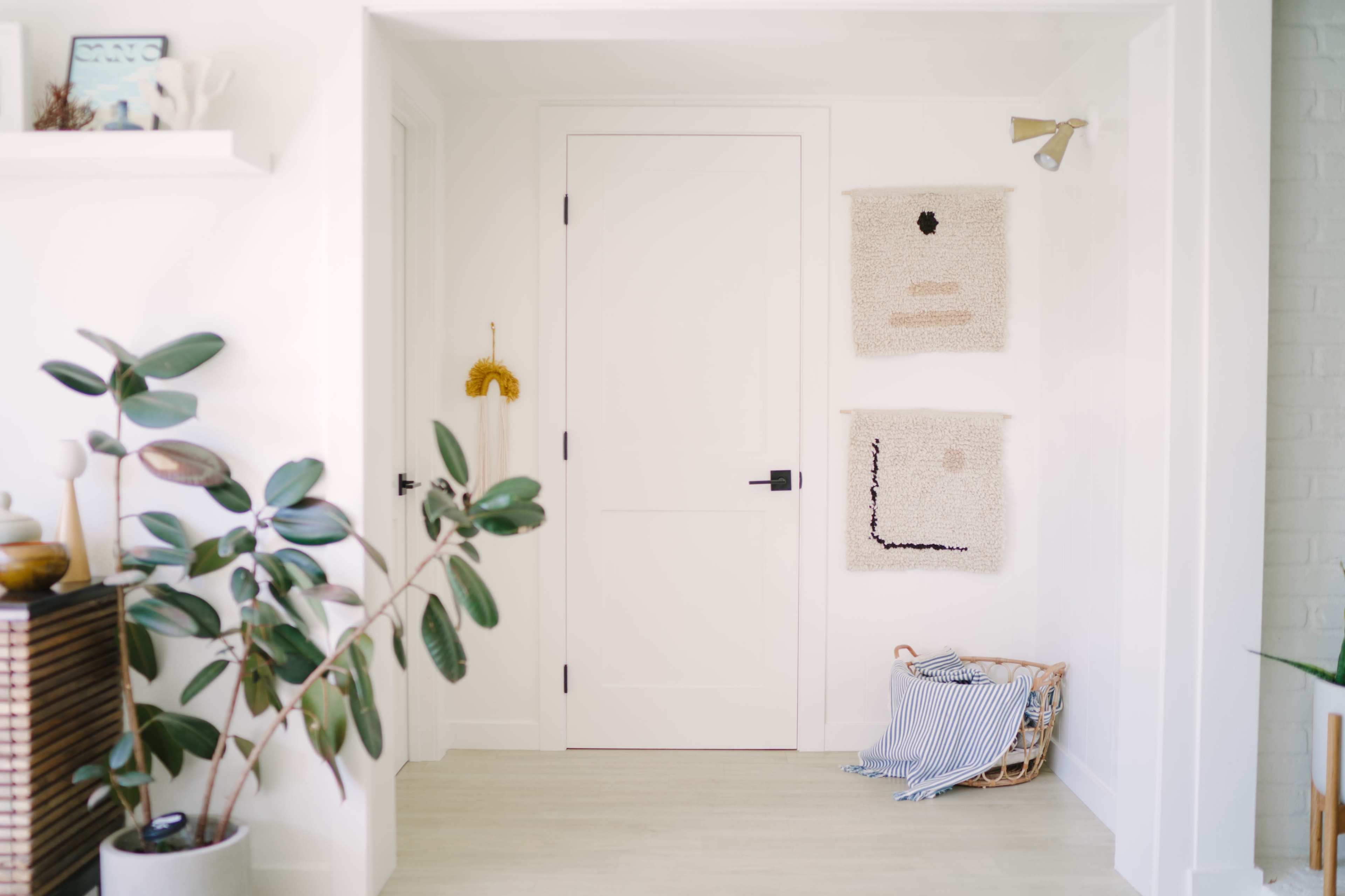 A bright hallway features a closed door, two textured wall hangings, and a plant next to a basket containing a folded striped blanket.
