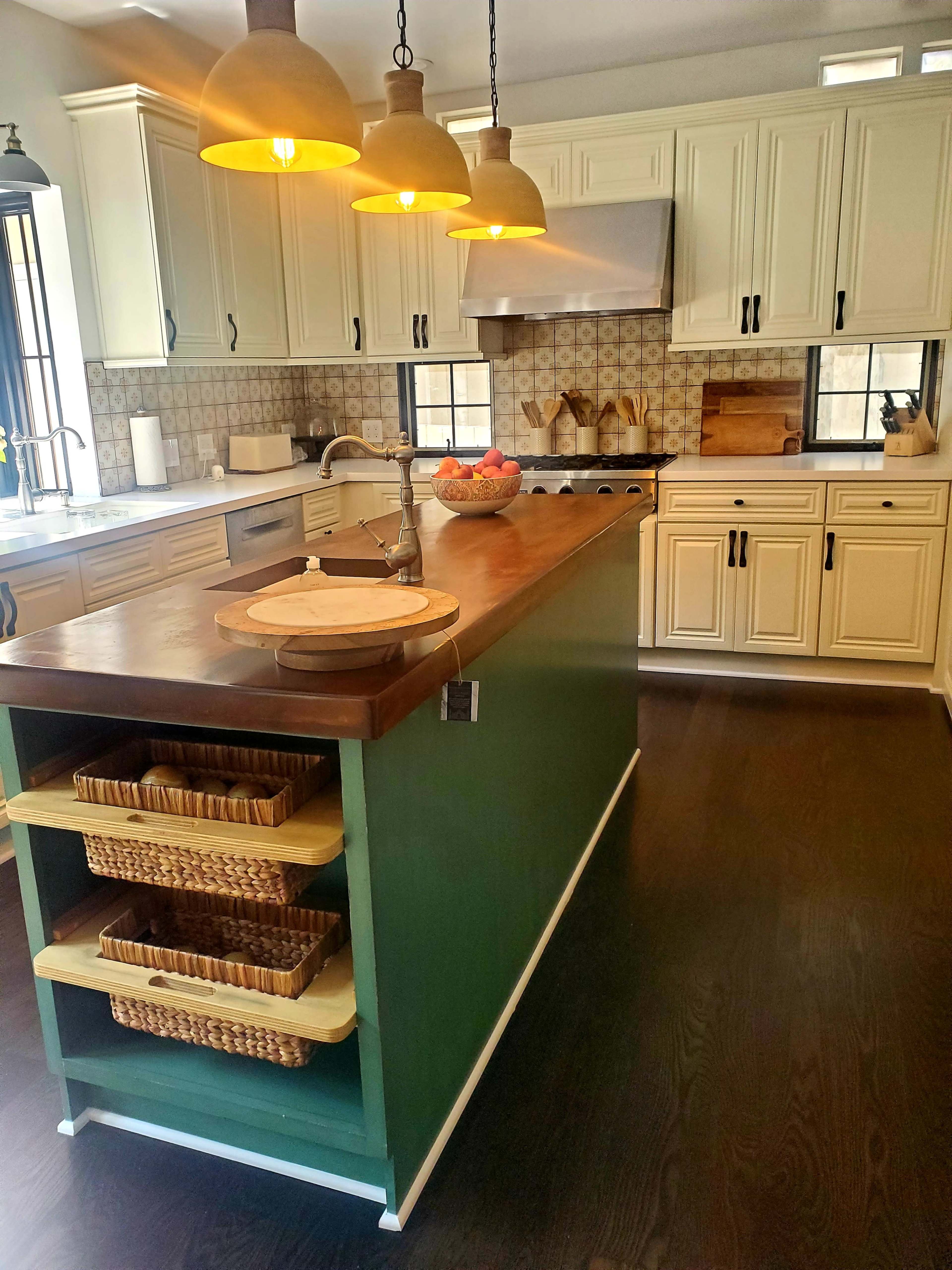 A kitchen features a central island with a wooden countertop and basket storage, surrounded by cream-colored cabinets and pendant lighting.