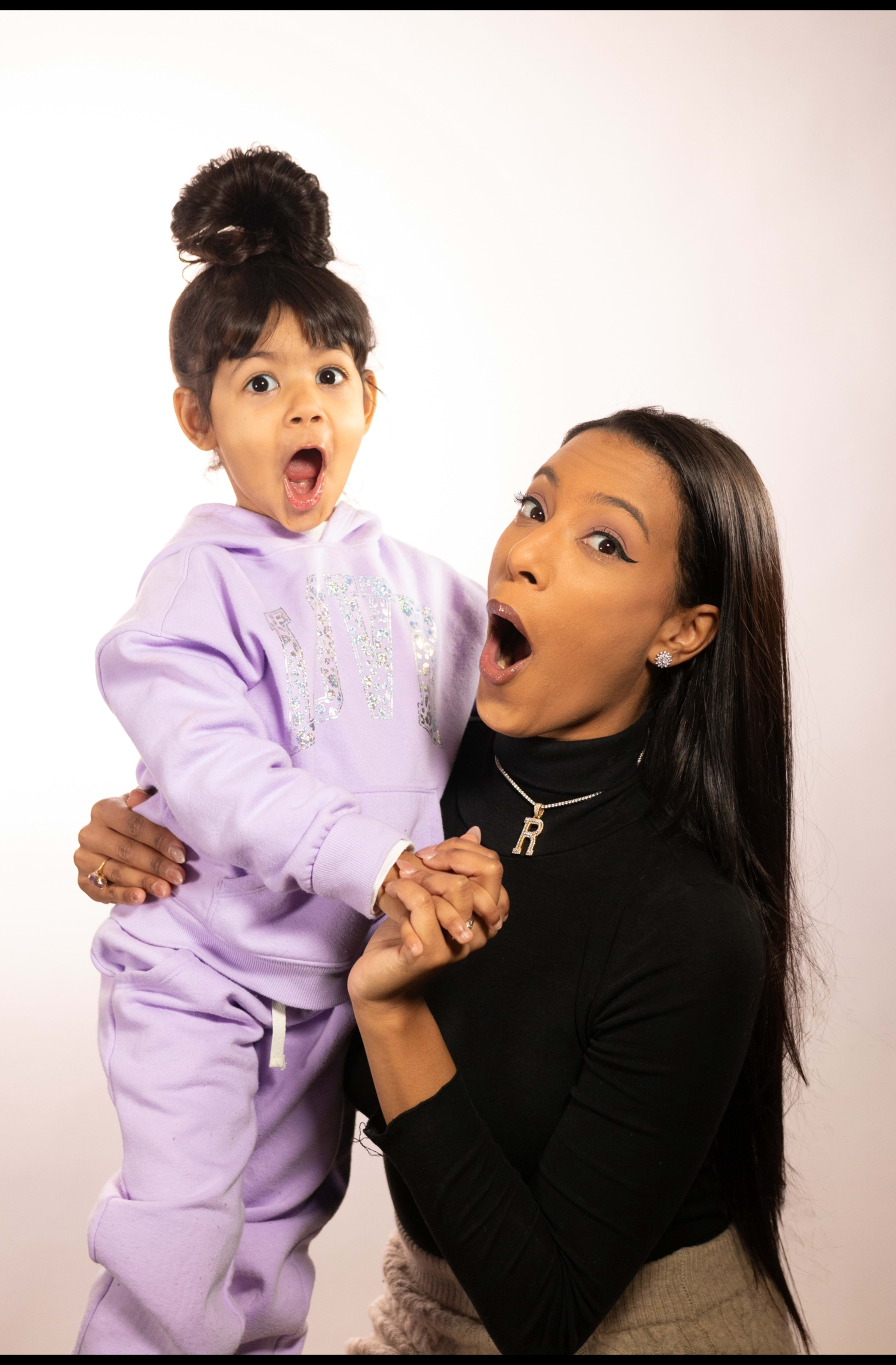 A woman and a child are both smiling and making surprised expressions while holding hands against a light background.