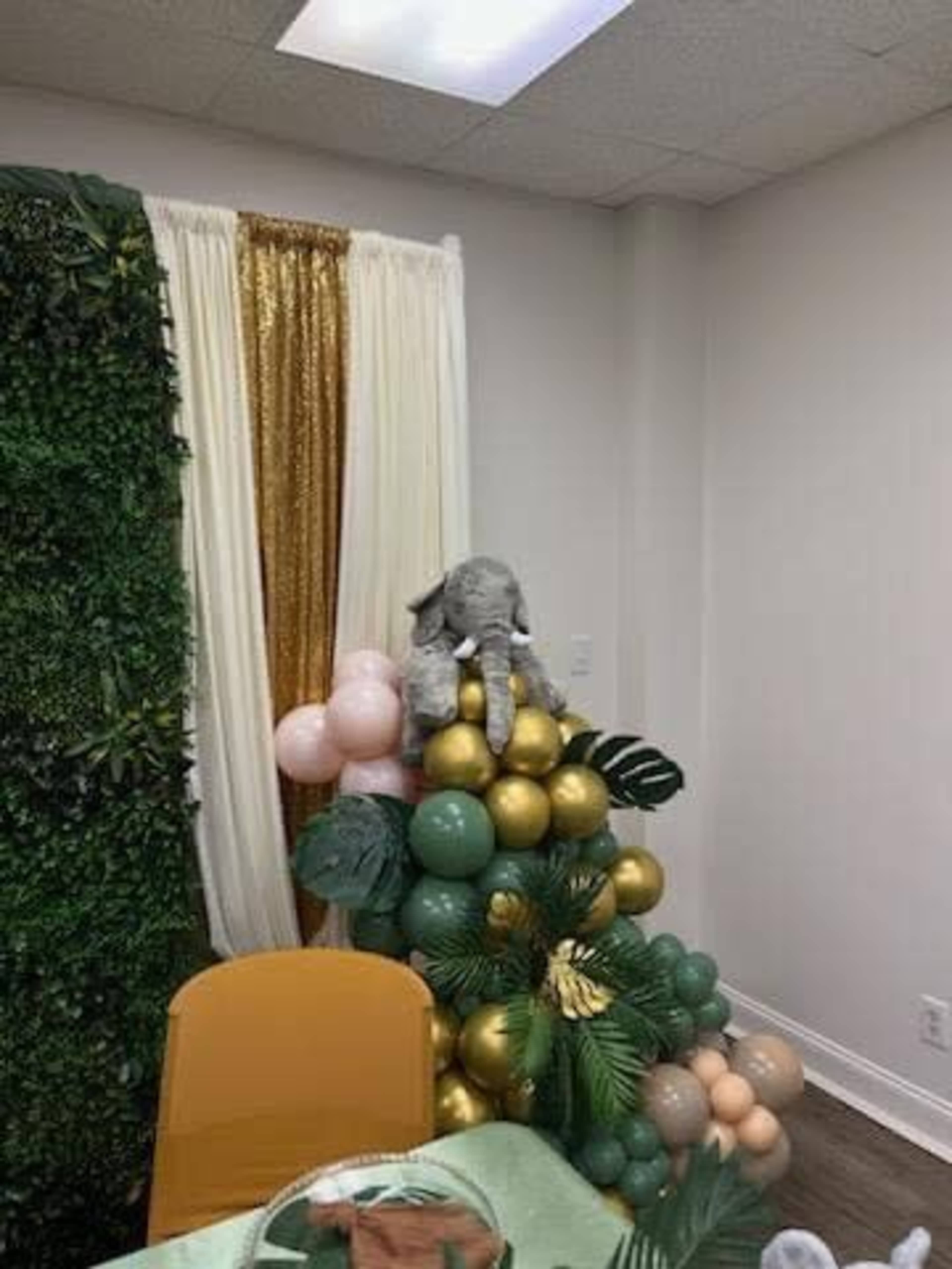 A stuffed elephant sits atop a decorative arrangement of green, gold, and pink balloons in a room with a leafy backdrop and a curtain.