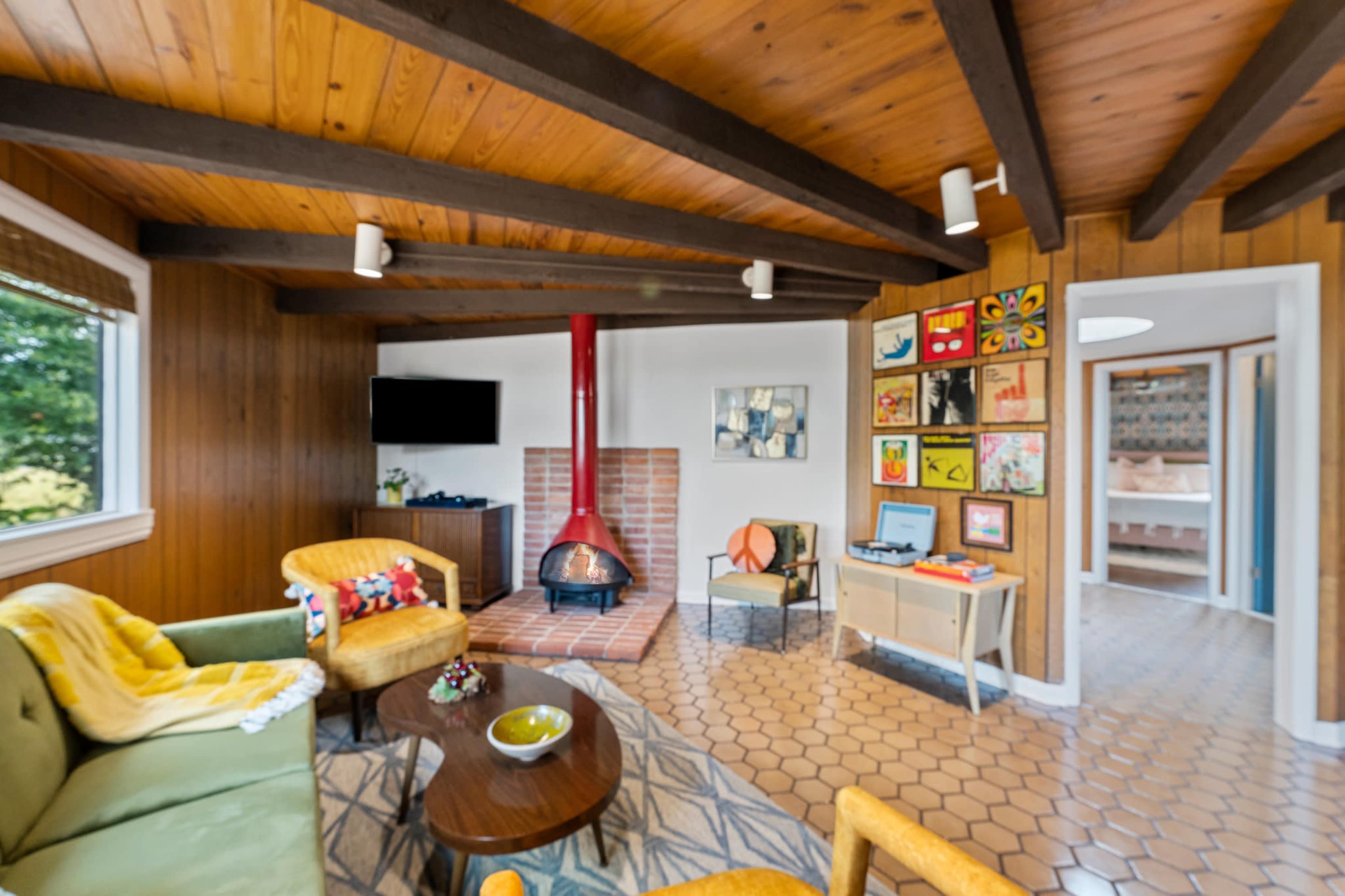 The image shows a cozy living room with a red fireplace, wooden beams, and a mix of artwork and furniture, including a green sofa and yellow armchairs.