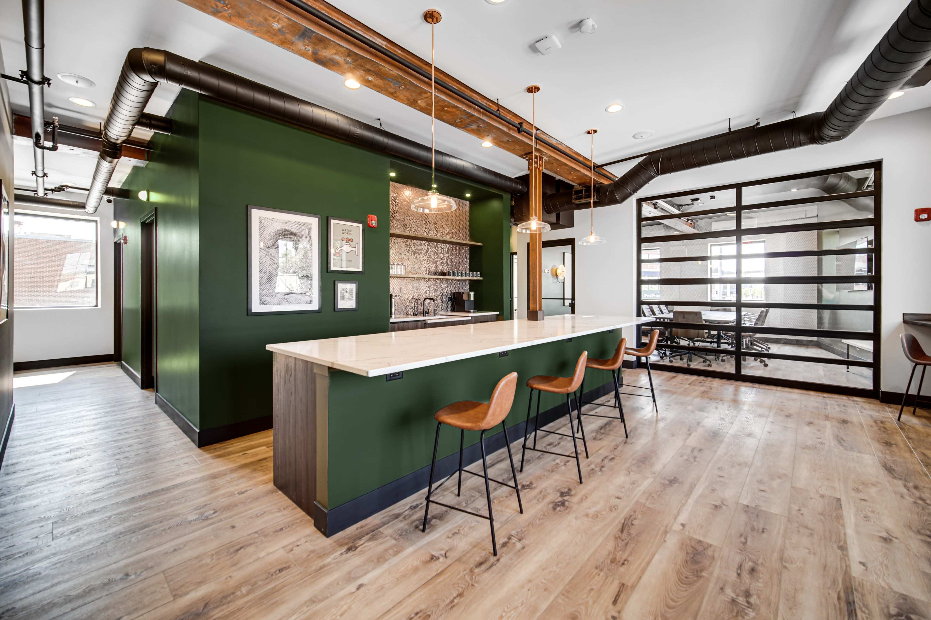 A modern kitchen area with a long countertop and wooden bar stools, featuring green walls and exposed ceiling beams.