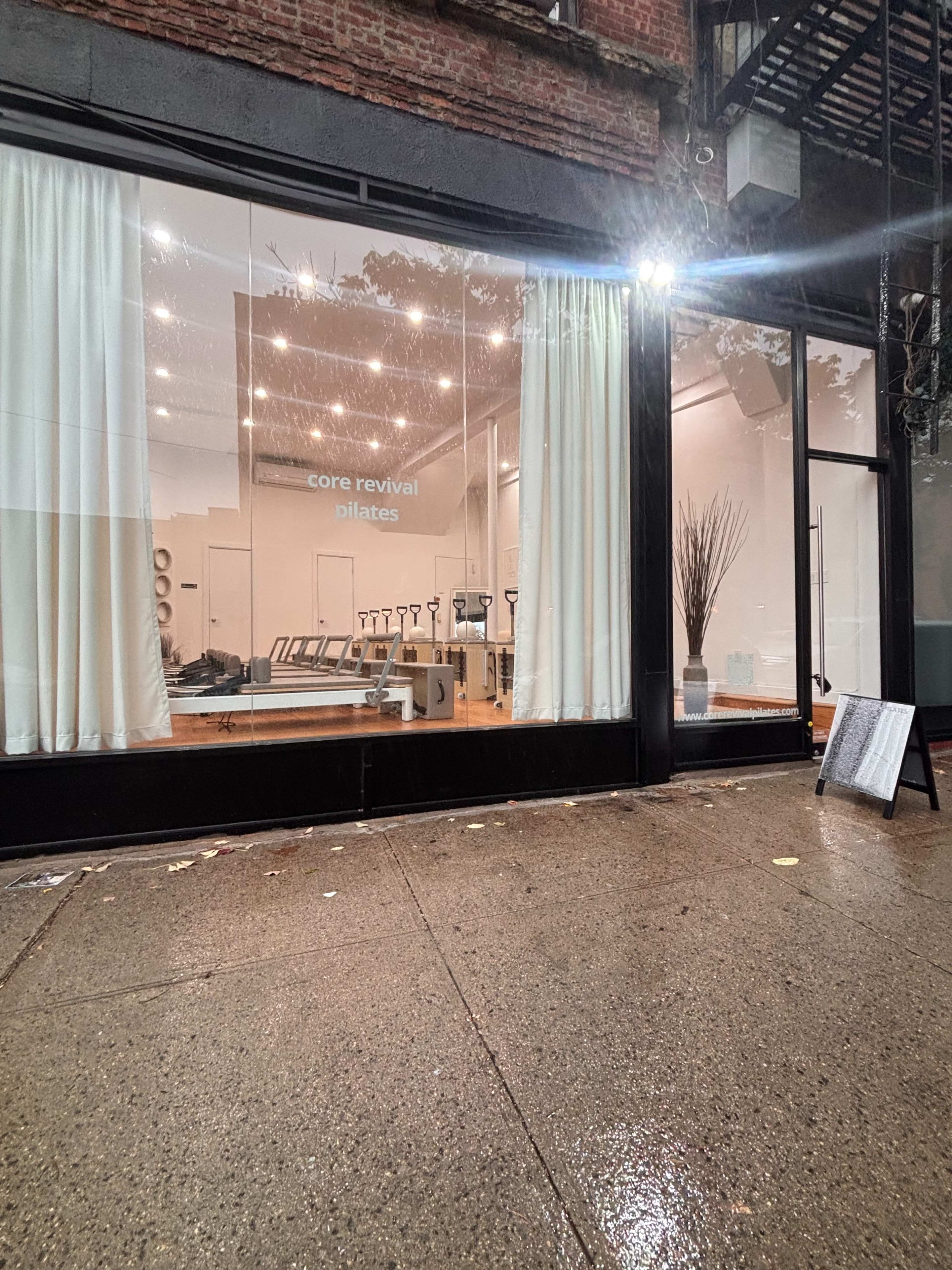 A Pilates studio is visible through the large front window, featuring exercise equipment and minimalistic decor, while rainwater collects on the sidewalk outside.