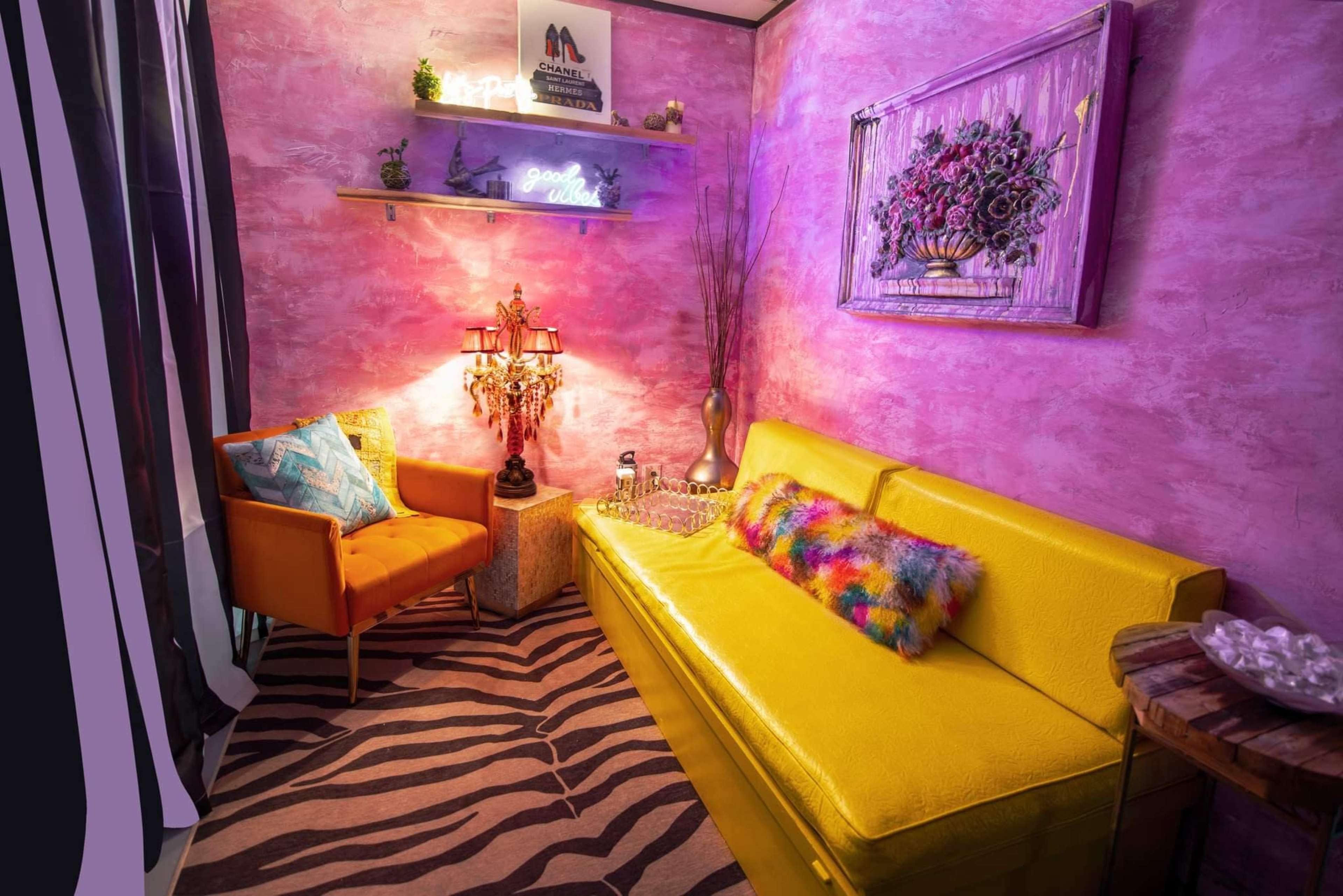A small, colorful interior space featuring a yellow sofa, an orange armchair, a zebra-striped rug, and decorative elements like a lamp and neon signs.