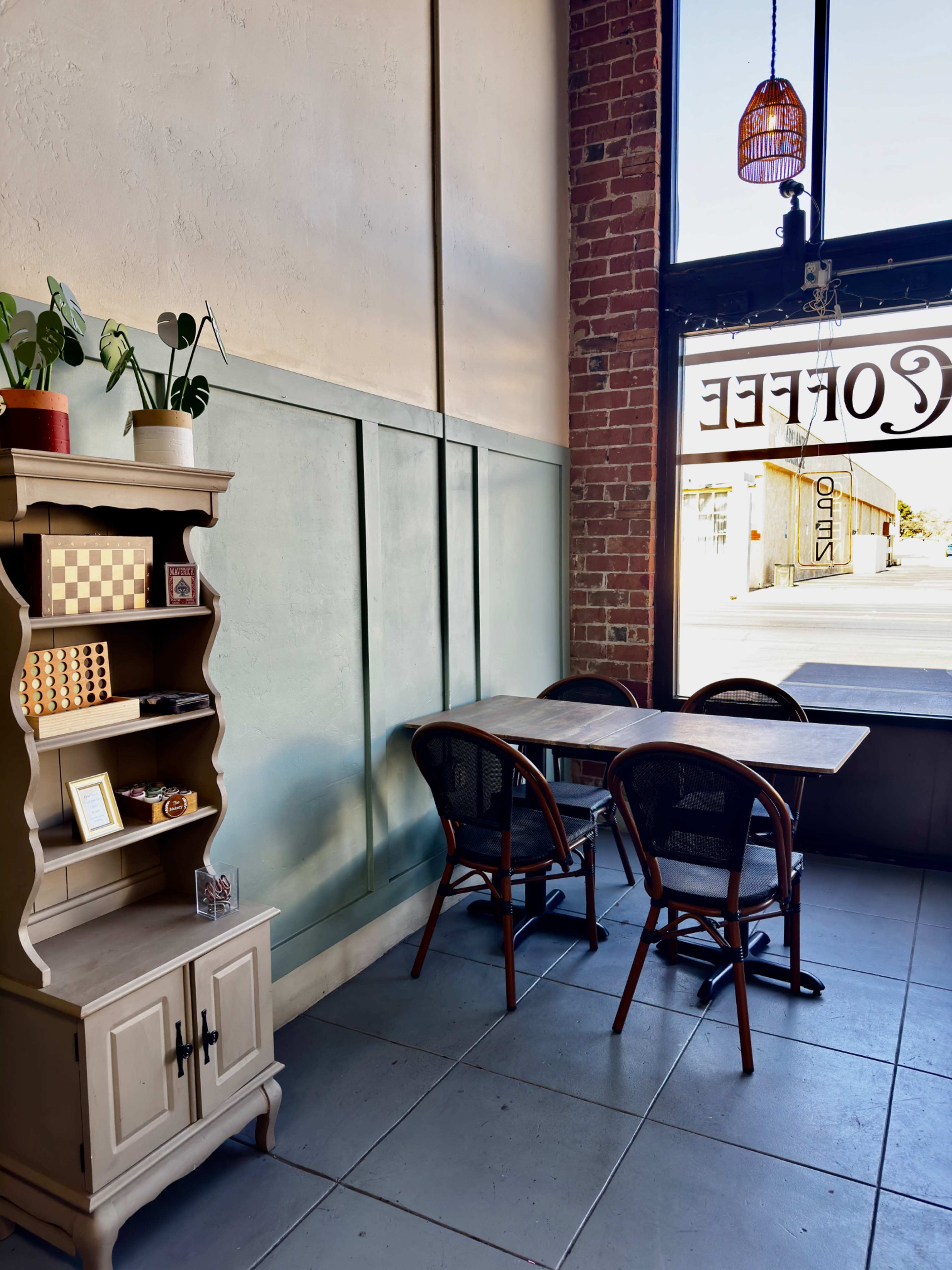 The image shows a cozy corner of a café featuring a small wooden bookshelf, two tables, and chairs near a large window with a "Coffee" sign.