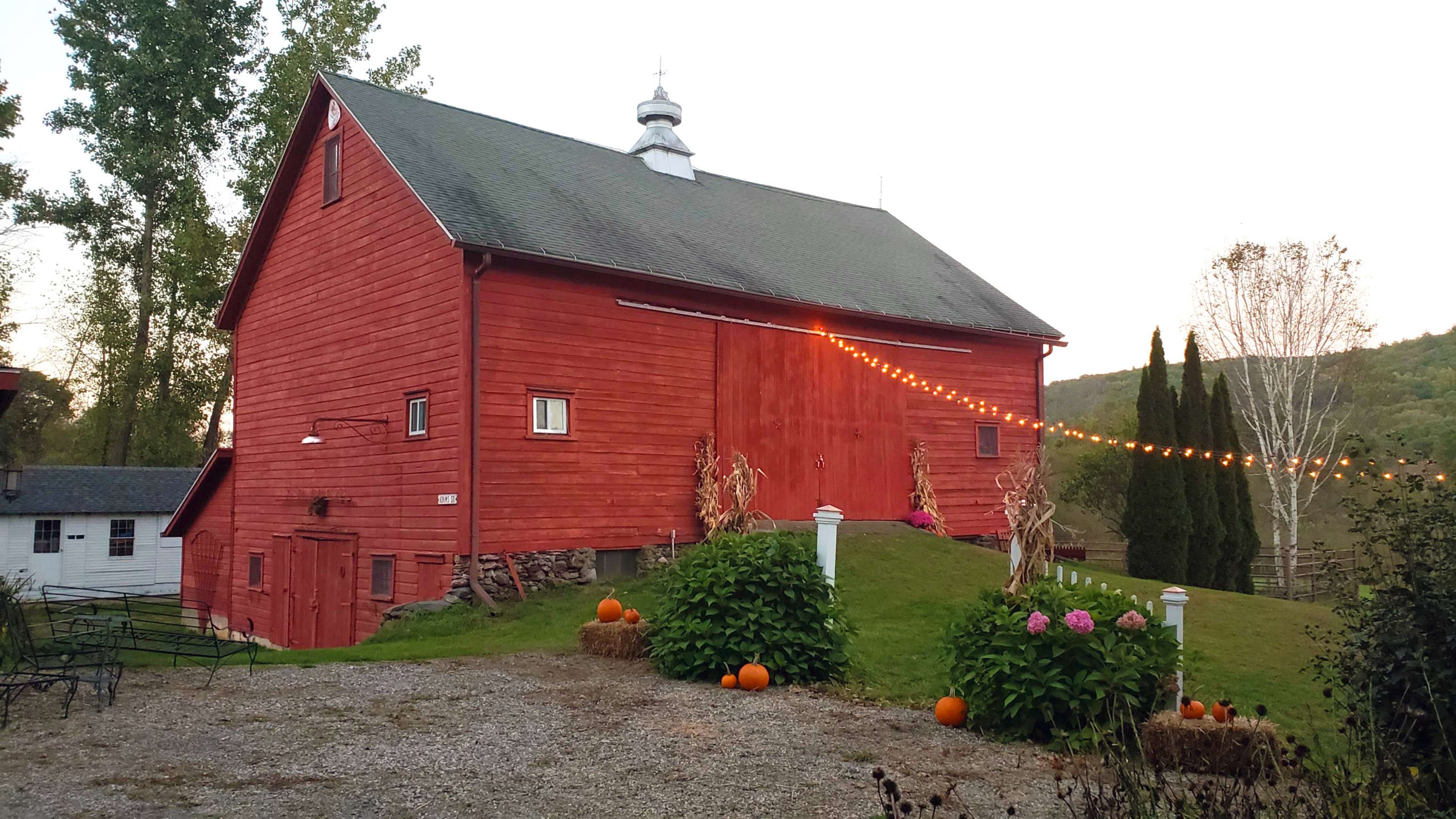 A red barn with decorative lights and pumpkins is set against a backdrop of trees and a hillside.
