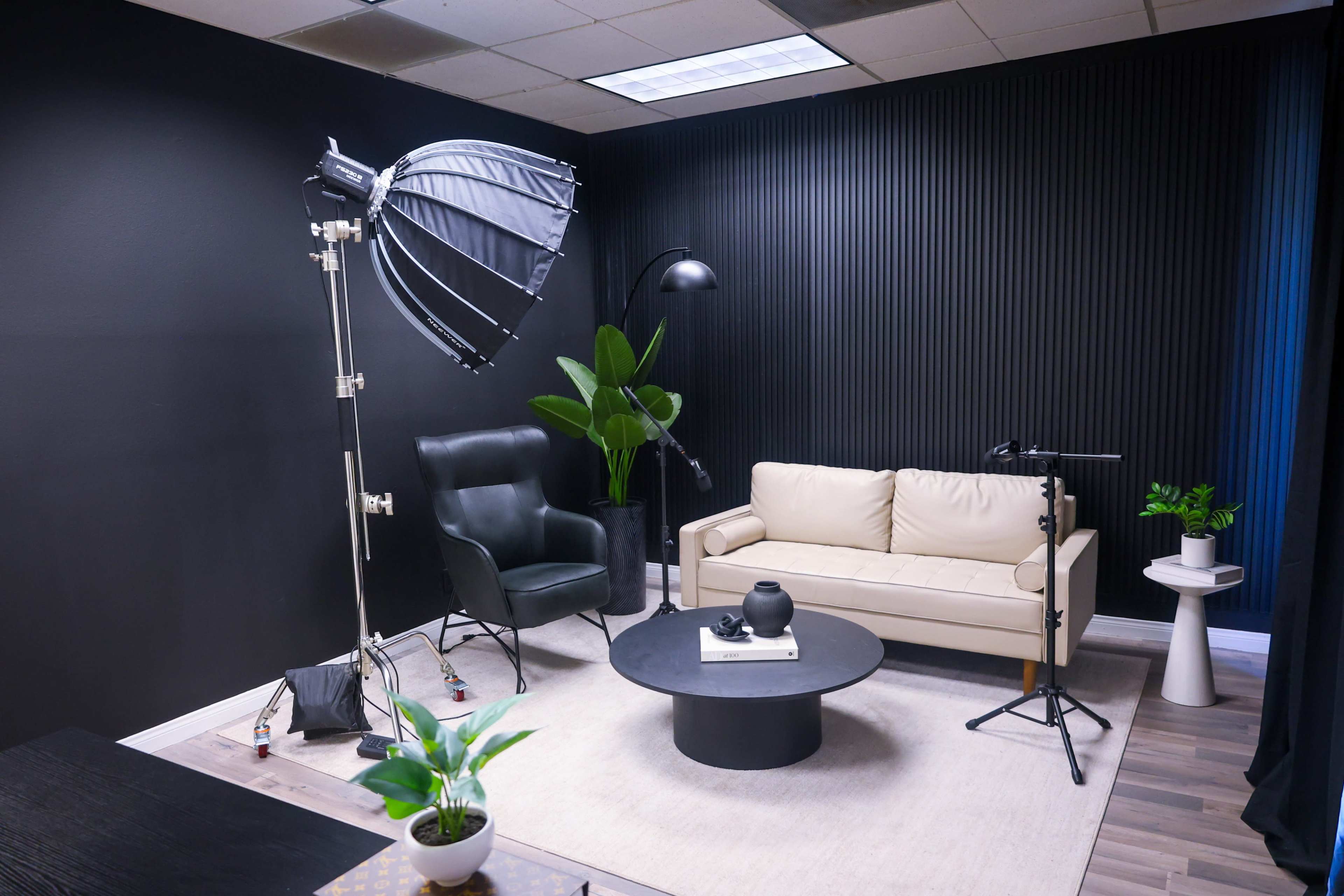 The image shows a modern studio space featuring a beige couch, a black chair, a round coffee table, and various lighting equipment against a dark wall.
