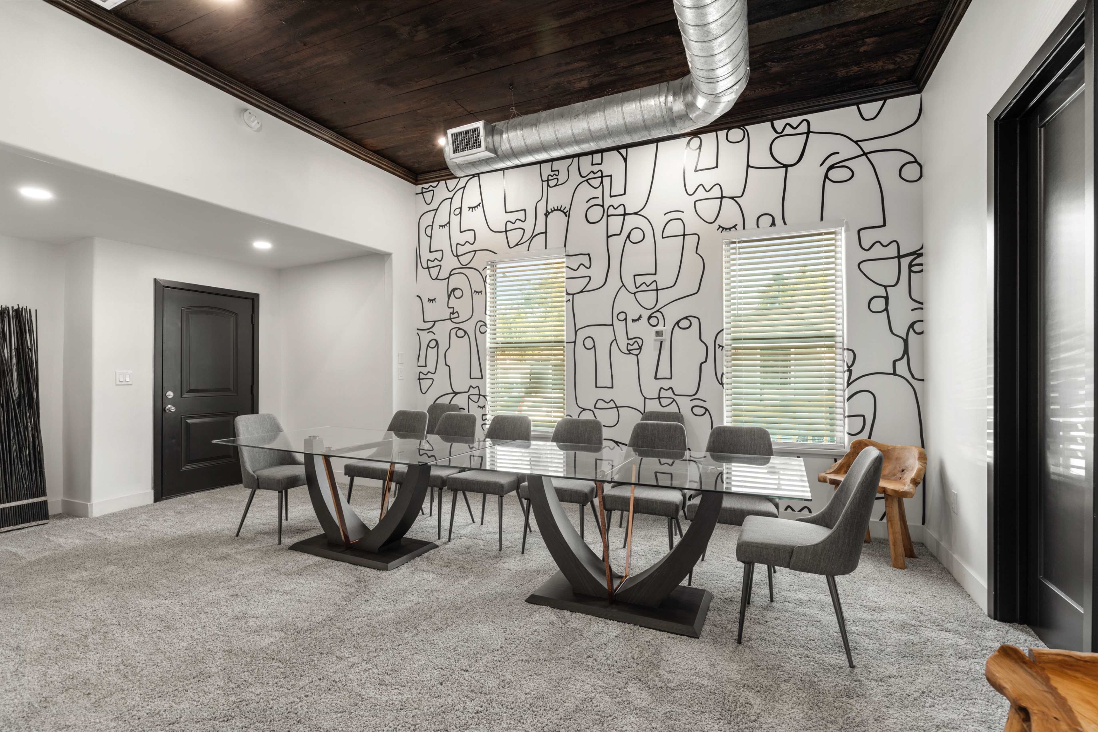A modern conference room features a large glass table surrounded by gray chairs, with a mural of abstract line art on the wall and a wooden accent chair in the corner.