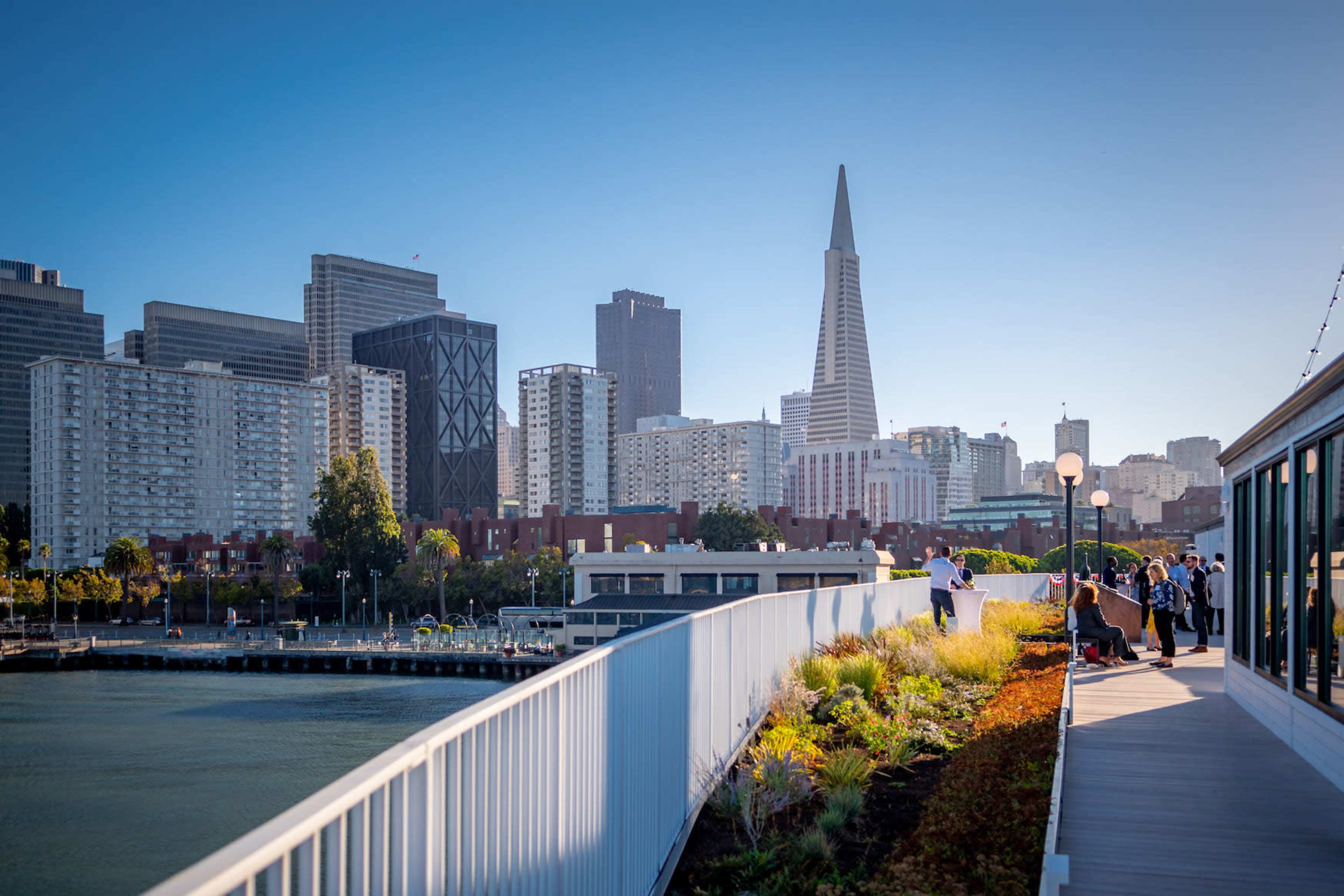 A waterfront walkway in a city with modern buildings, including a prominent spire, along a calm body of water.