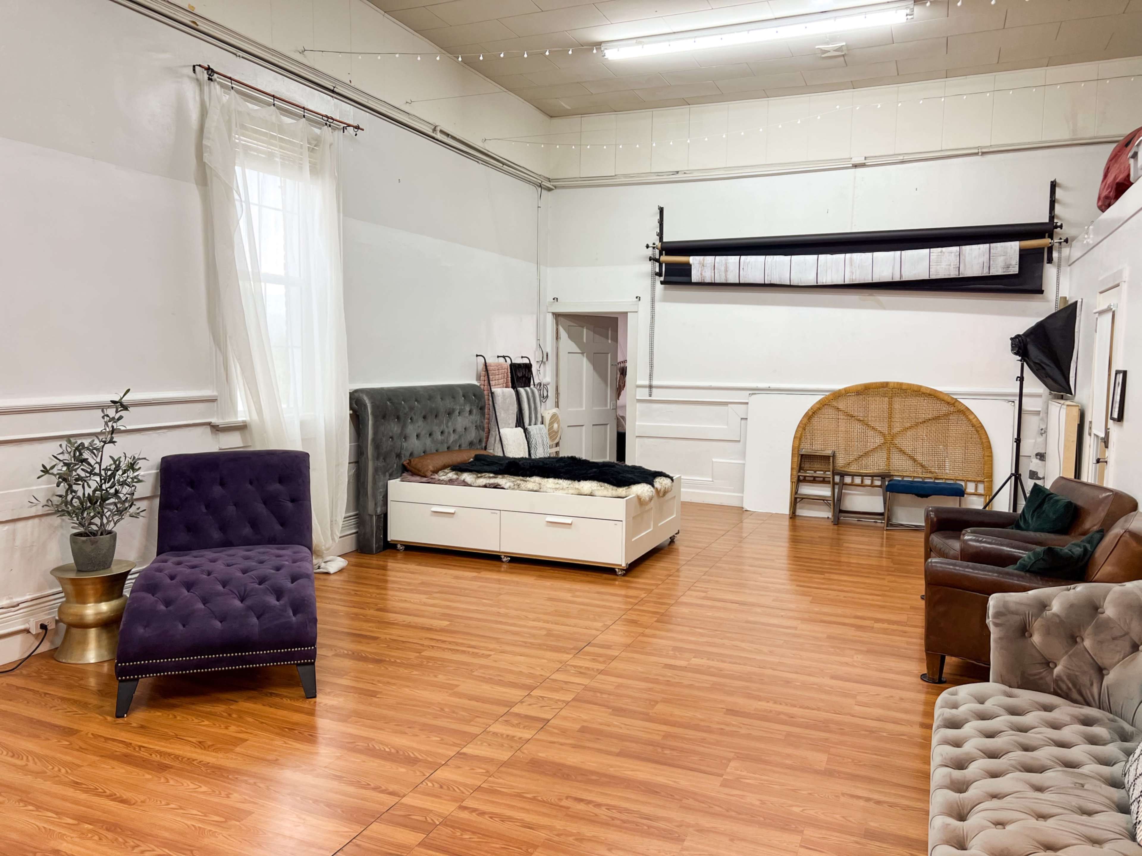 The image shows a spacious room with wooden flooring, featuring a bed with a black fur throw, a purple upholstered chair, a brown leather sofa, and a decorative plant in a pot.