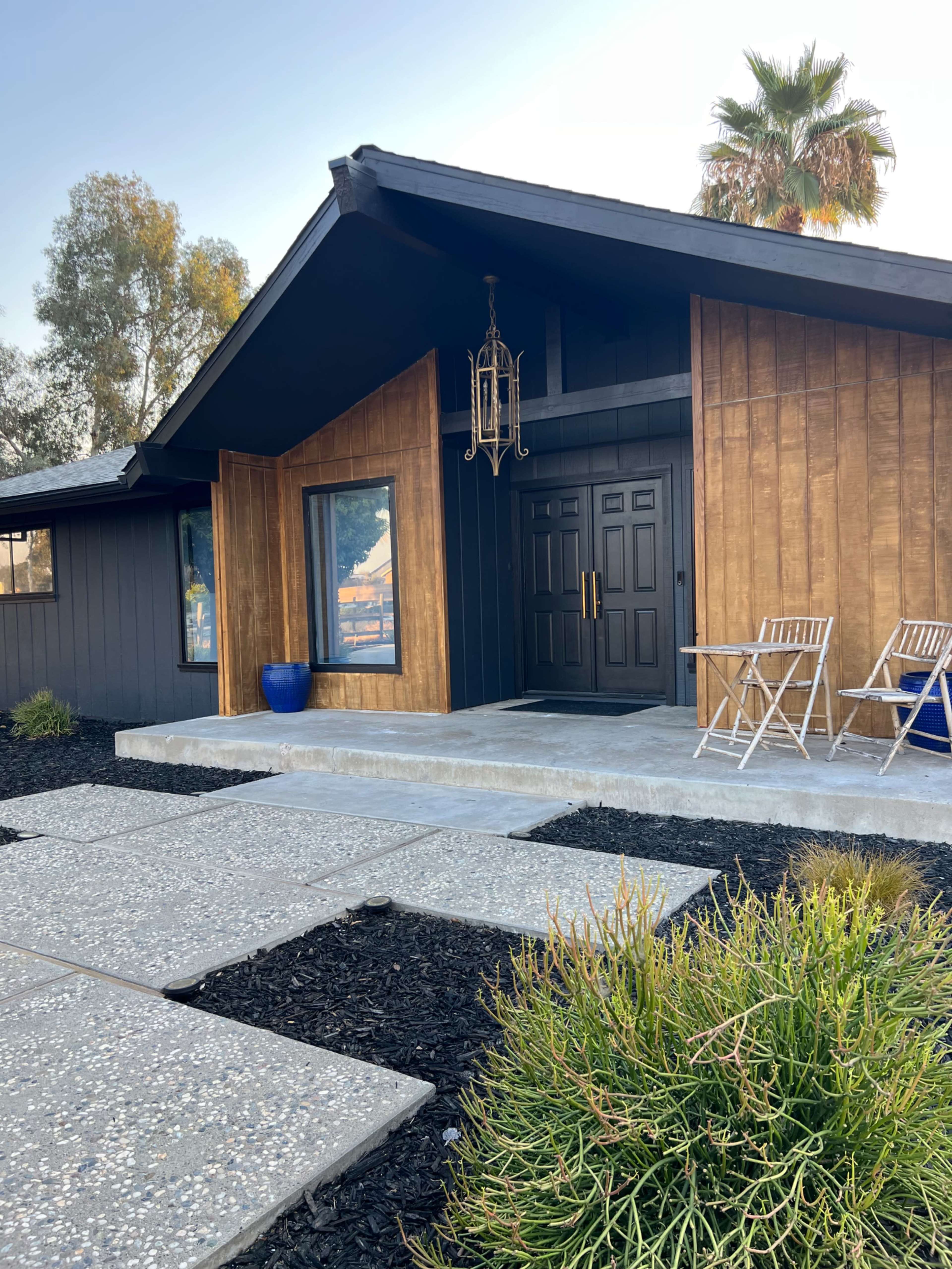 A modern single-story house features a prominent entrance with double doors, flanked by large windows and surrounded by a landscaped pathway and seating area.