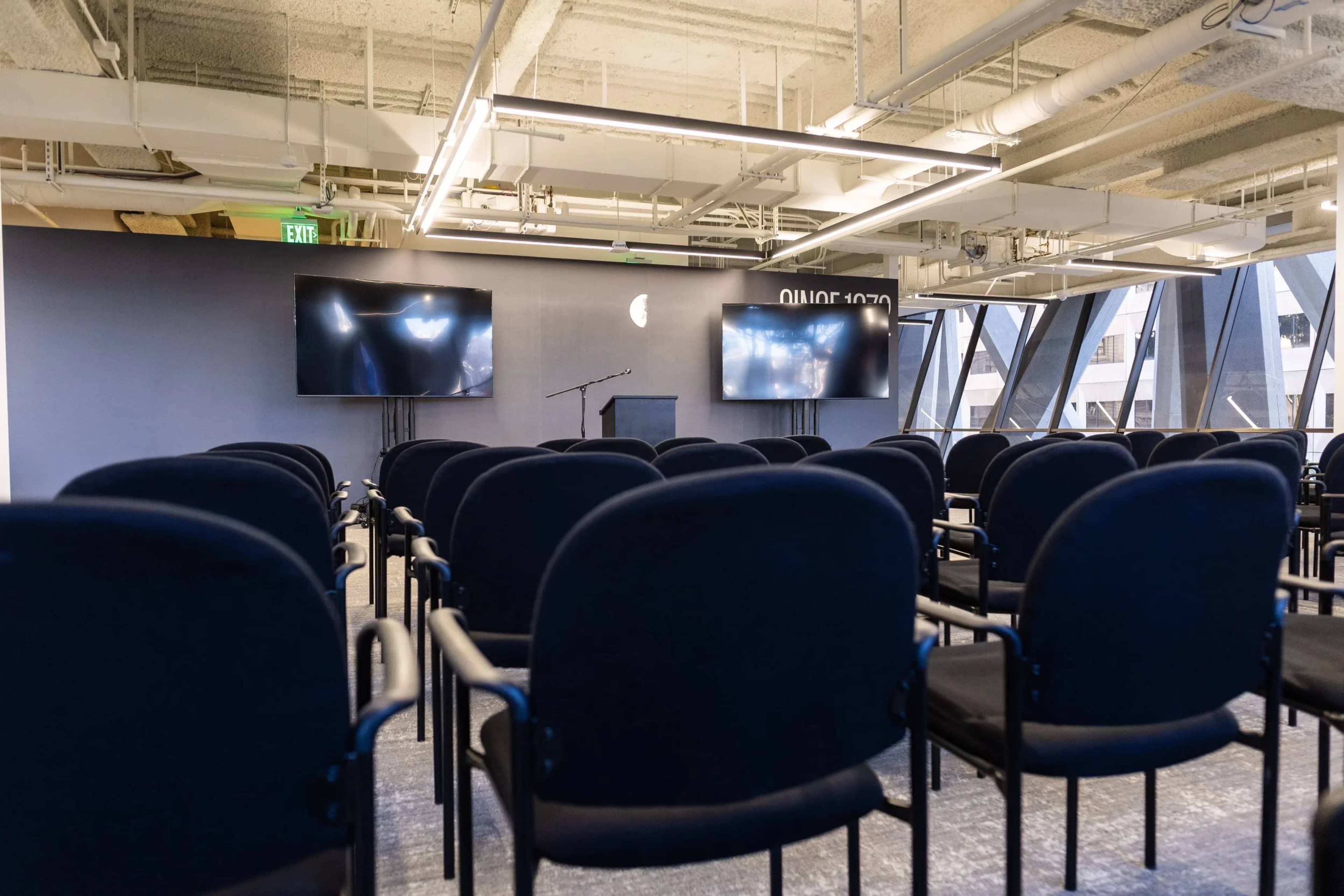 The image shows a conference room set up with rows of black chairs facing a podium and two large screens on the wall.