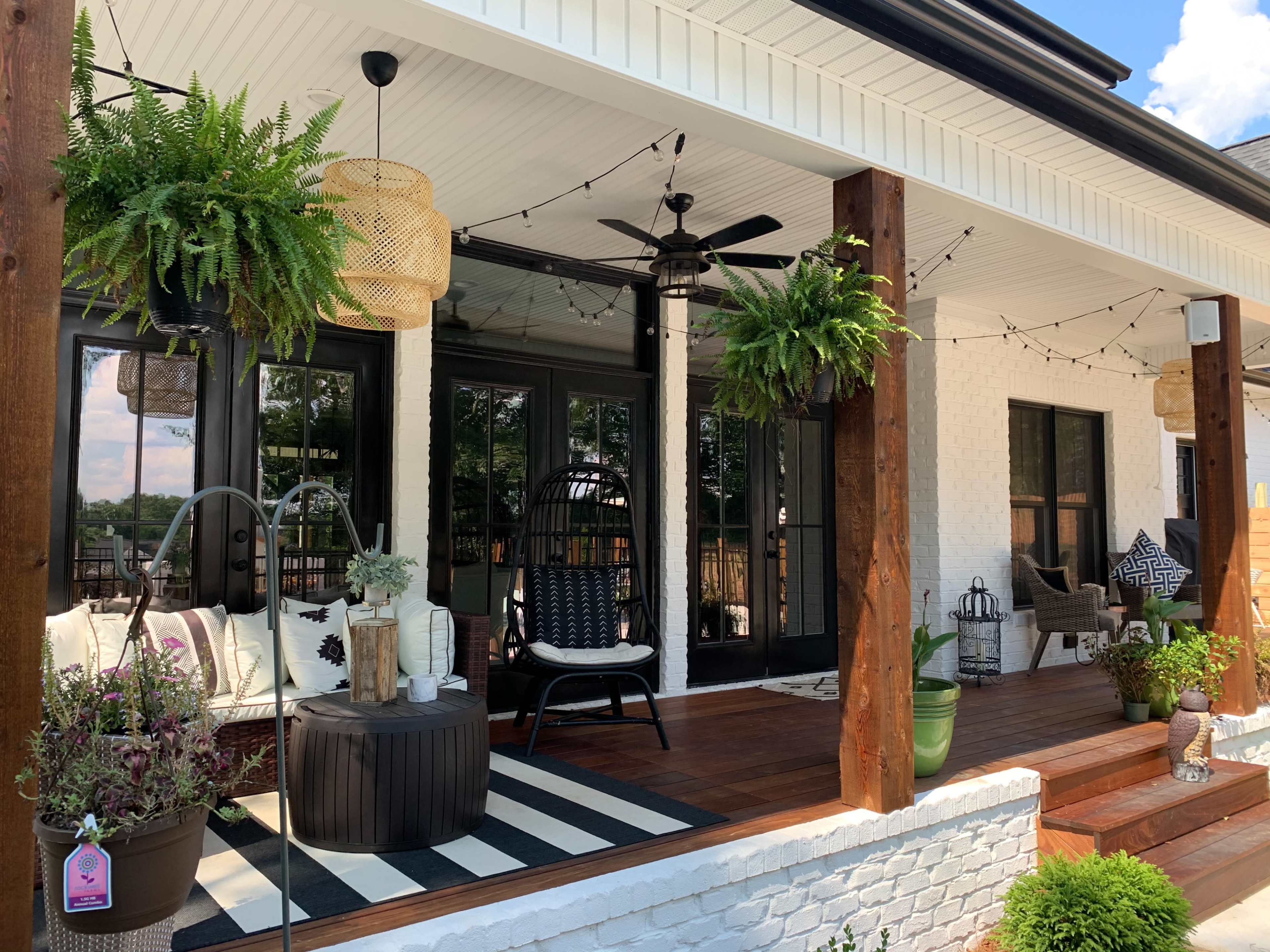 The image shows a covered porch area with comfortable seating, hanging plants, a coffee table, and decorative lighting.