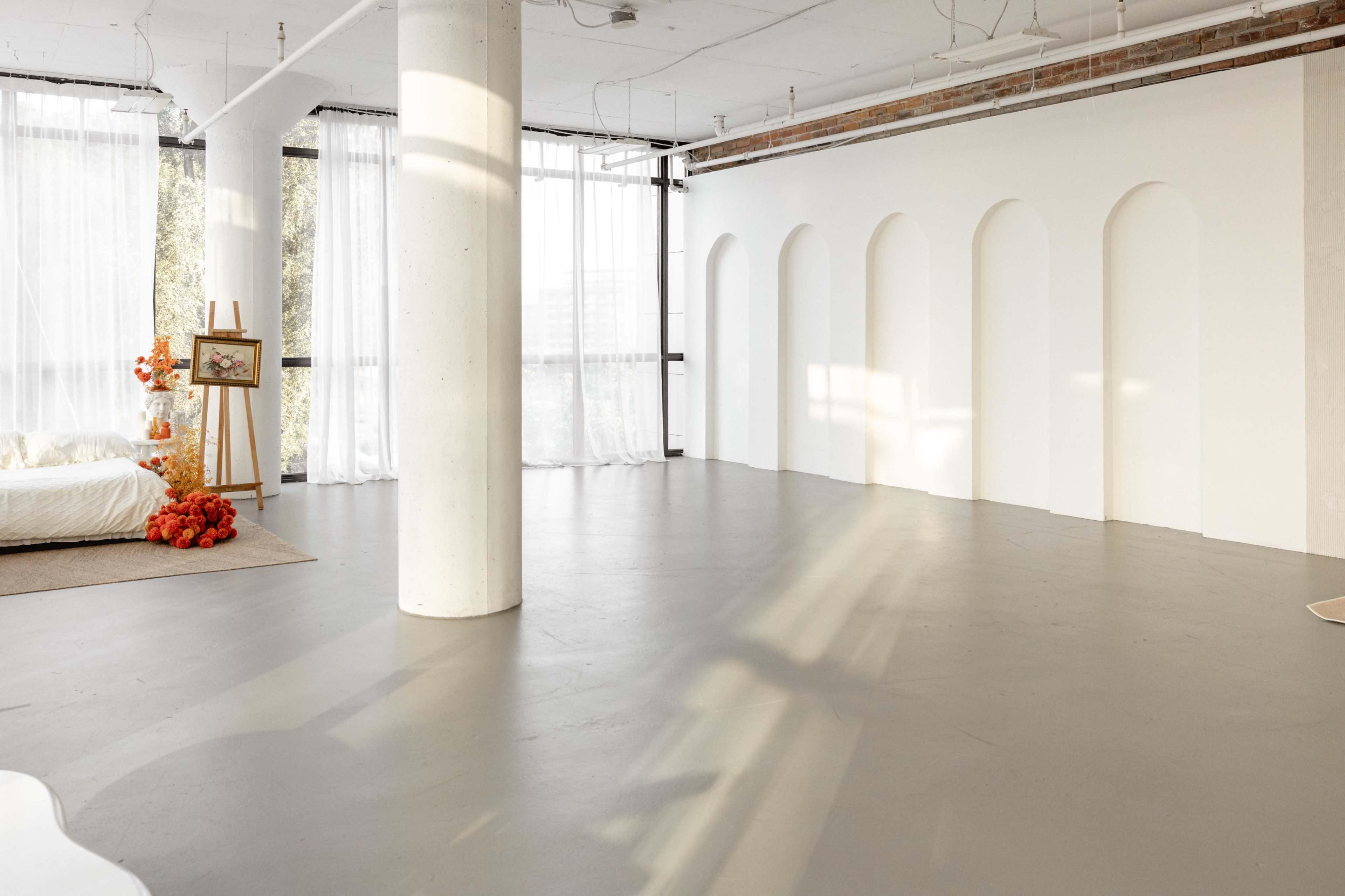 The image shows a spacious, well-lit interior with large windows, a column in the center, and a minimalist design featuring arched wall niches.
