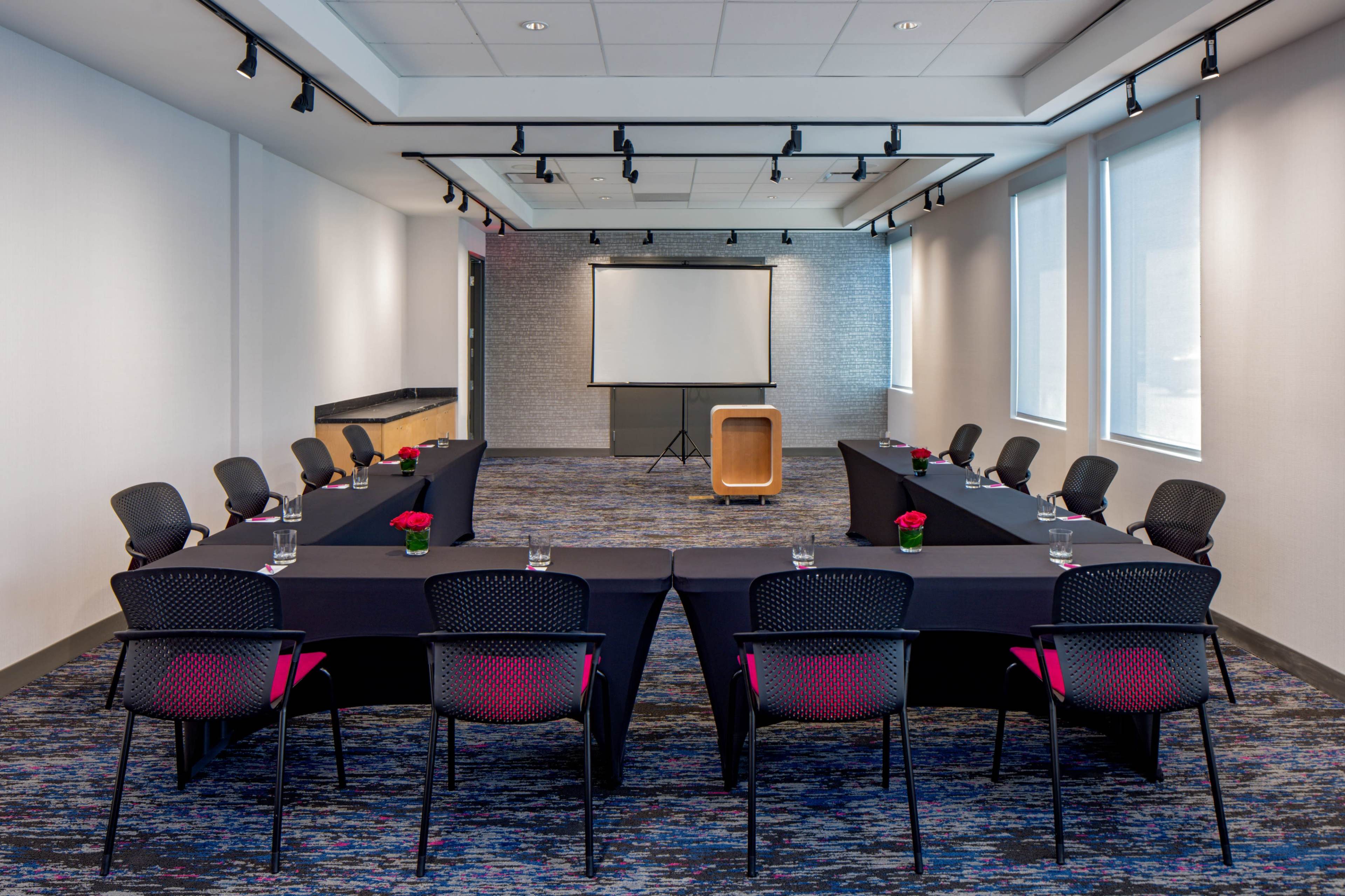A conference room is set up with a U-shaped arrangement of black tables and chairs, featuring a projector screen at one end and small flower arrangements on each table.