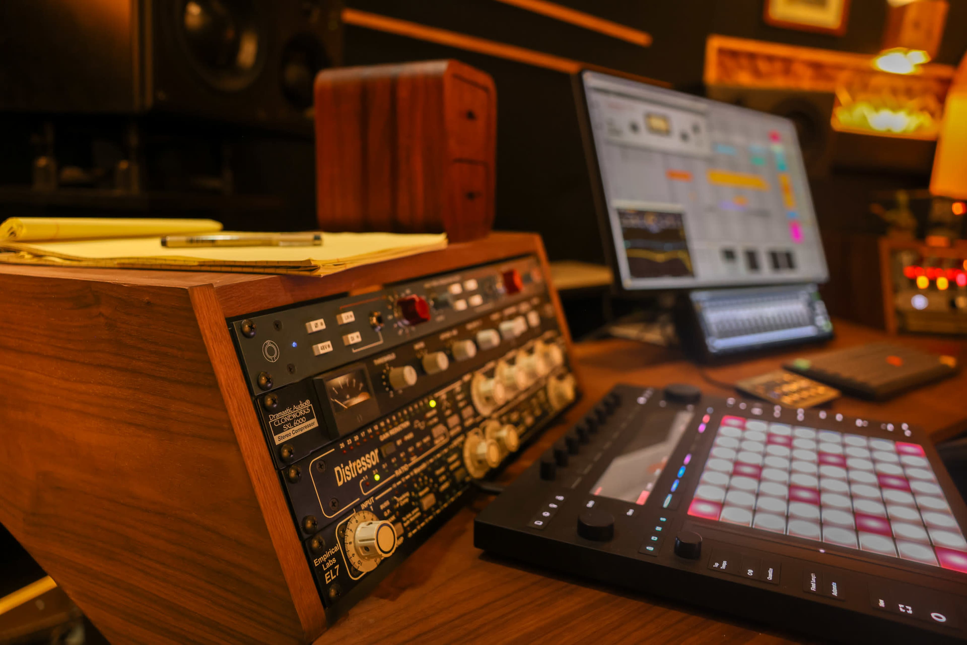 The image shows a close-up of a music production workstation featuring audio equipment, a digital audio workstation on a monitor, and a MIDI controller.