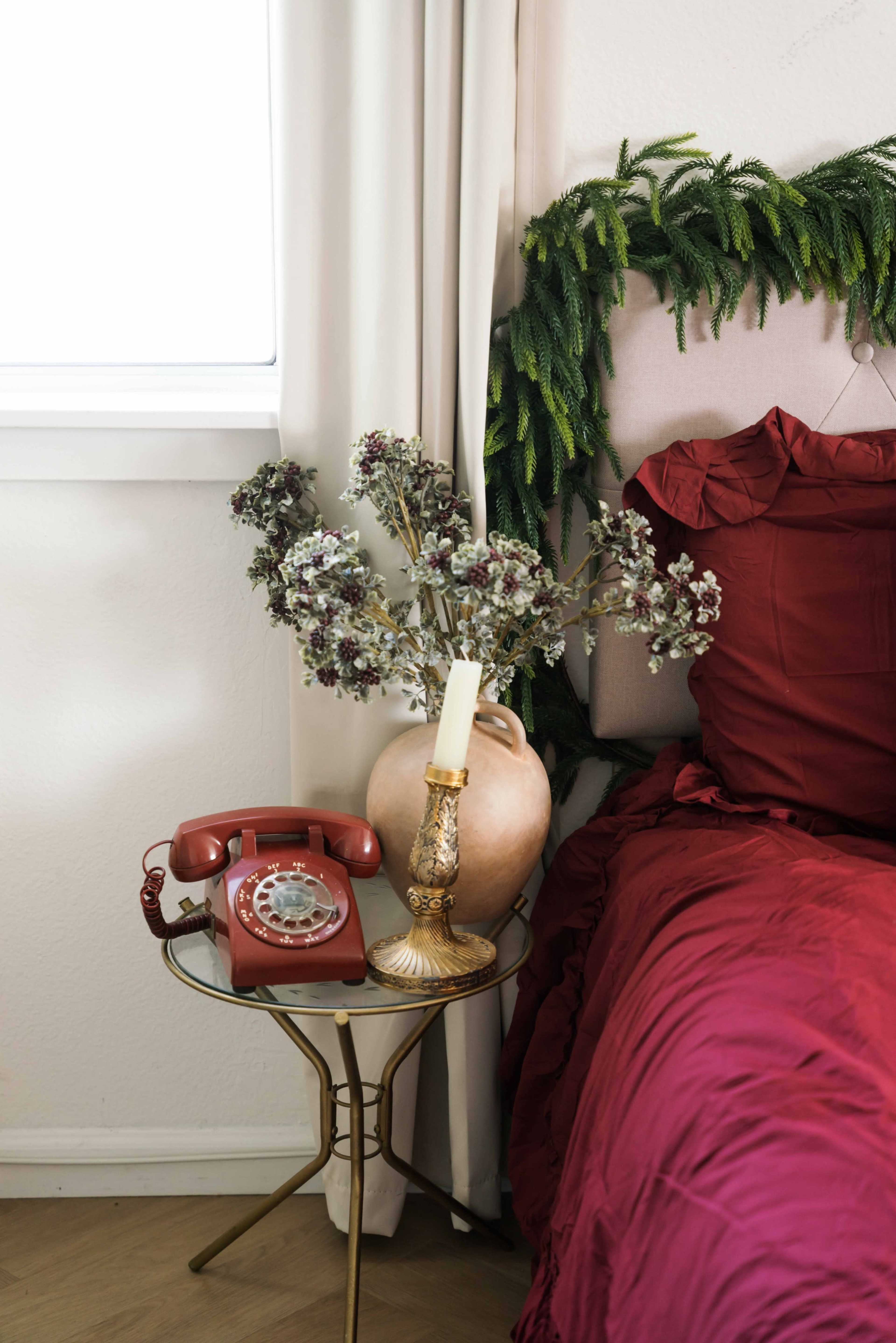 A red rotary phone sits on a small gold table beside a vase with dried flowers and a candle, near a bed with a rich red comforter.